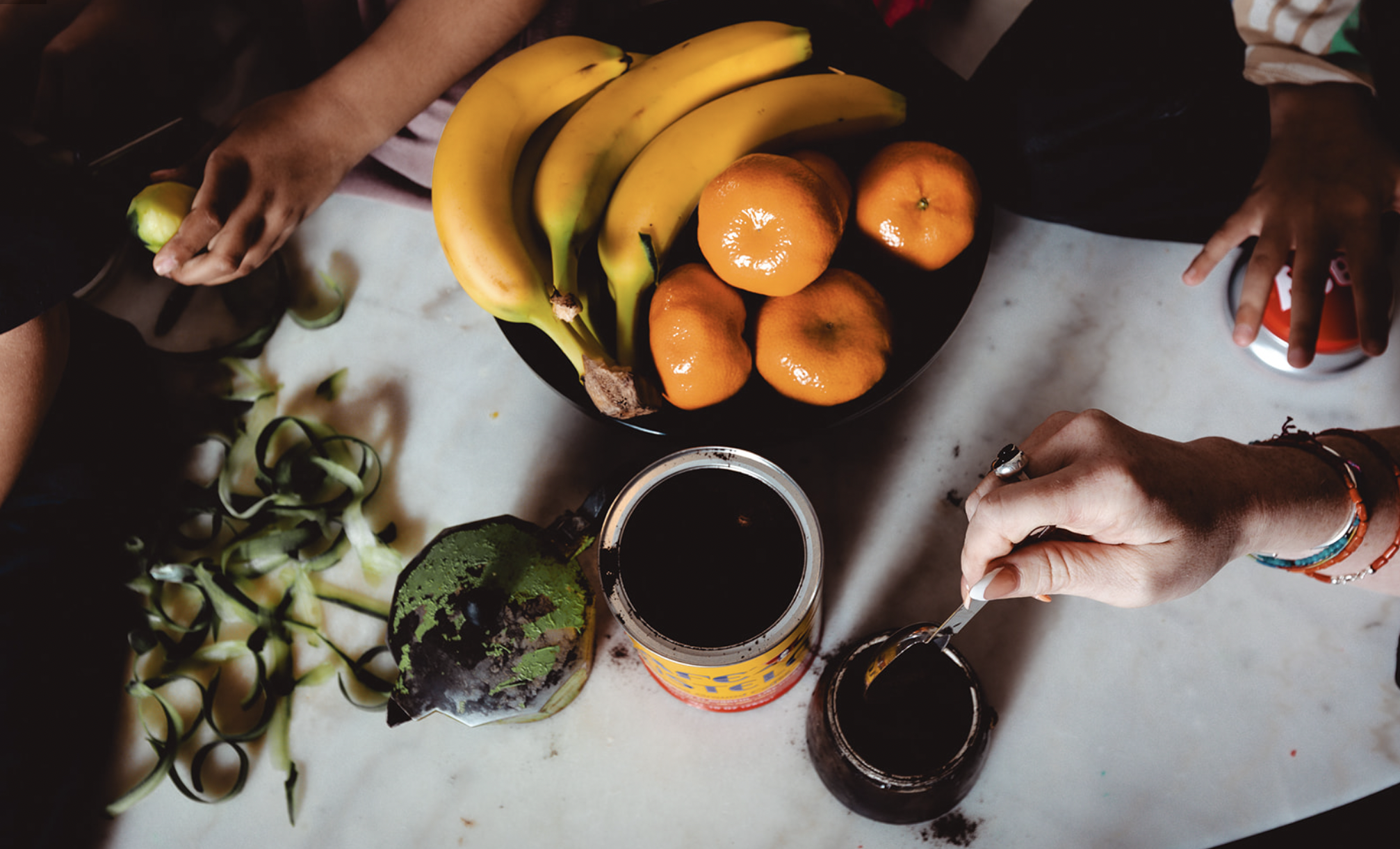 A top-down view of a table with a bunch of bananas, oranges, a jar of coffee, a cup of coffee, a container of tea, cilantro, and a person's hand holding a spoon.
