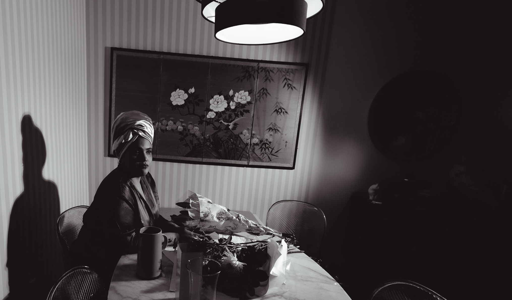 A woman with a headwrap sitting at a table with paper, a vase, and a mug, in a room with striped wallpaper and Asian art on the wall, with a dark shadow on the right side of the image.