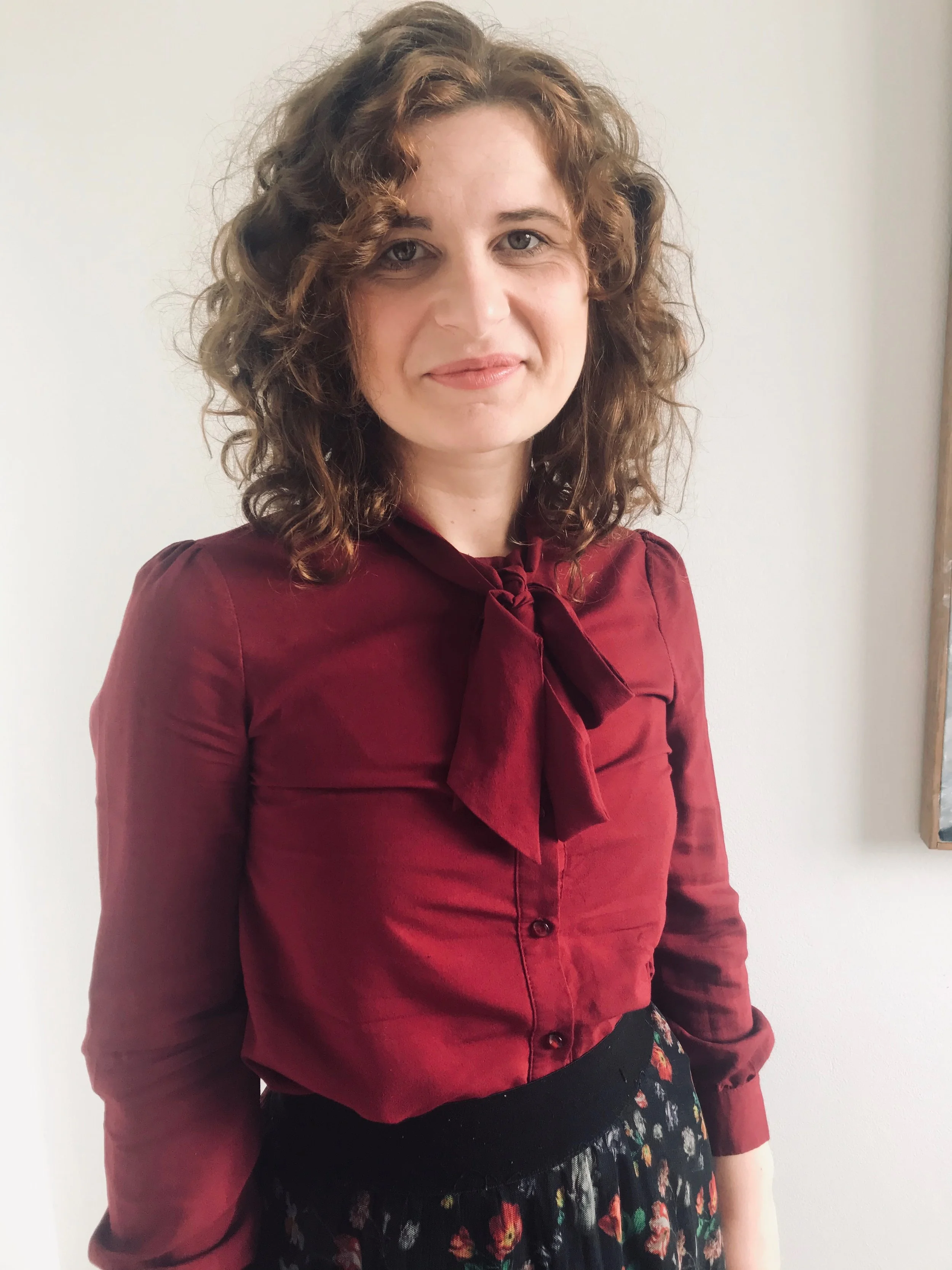 A woman with curly brown hair wearing a maroon blouse with a bow and a black skirt with a floral pattern standing against a plain white wall.