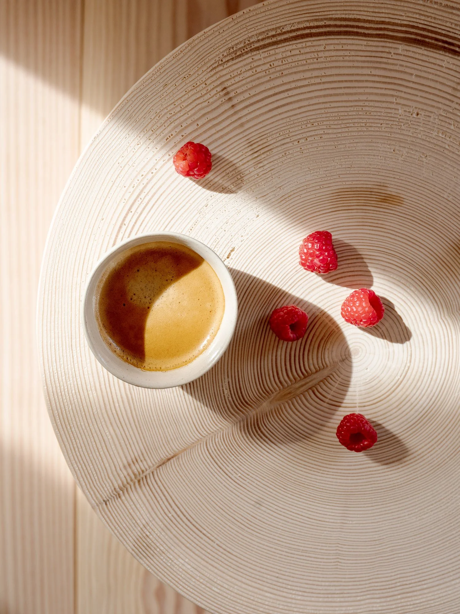 Ein weißer Kaffeeteller mit Kaffee und roten Himbeeren, auf einem runden Holztablett.