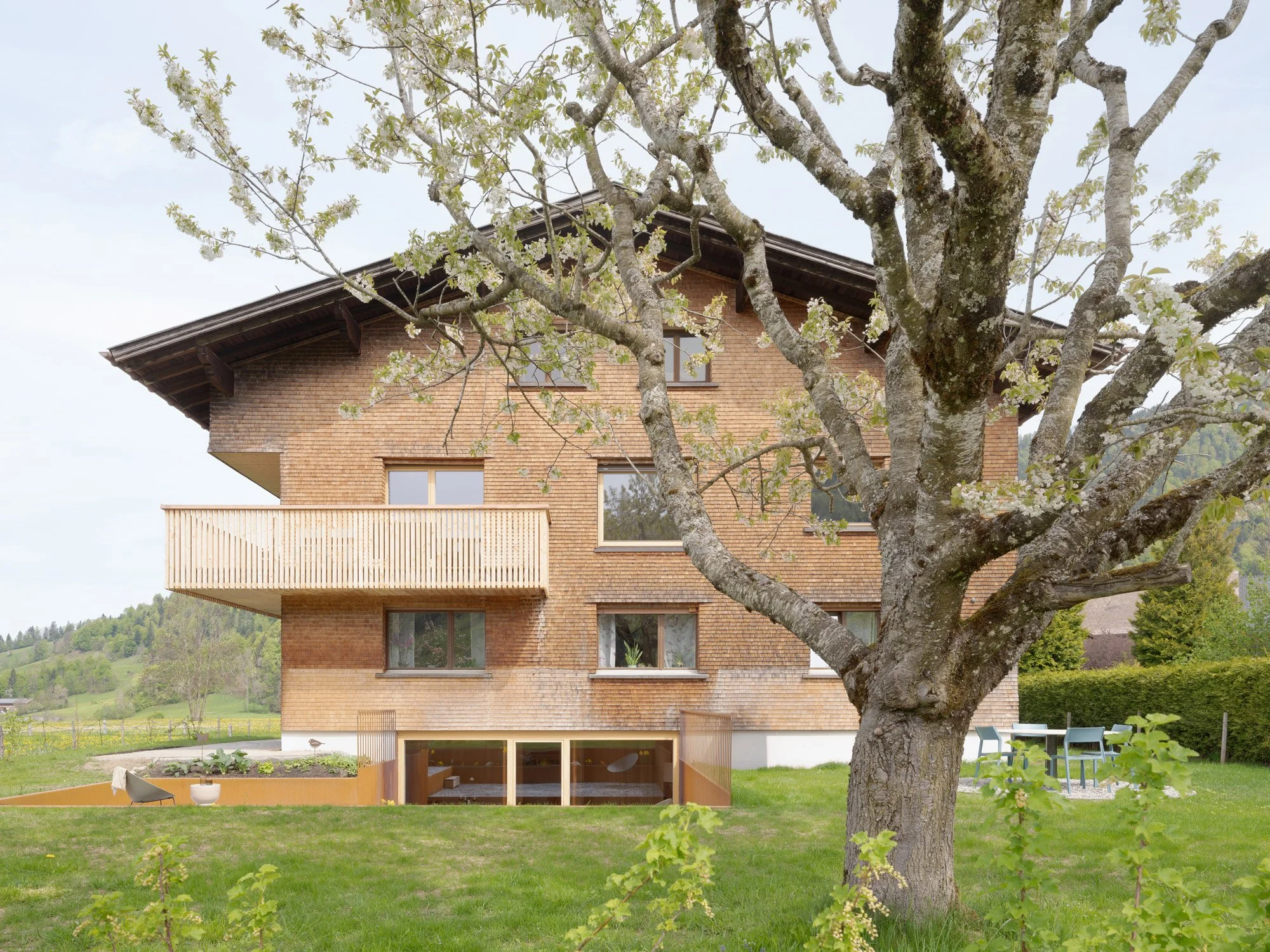 Mehrstöckiges Haus mit Holzalufen und einem Baum im Vordergrund, im Garten sind Stühle und Tische sichtbar, umgeben von grüner Wiese und Bäumen in einer ländlichen Gegend.