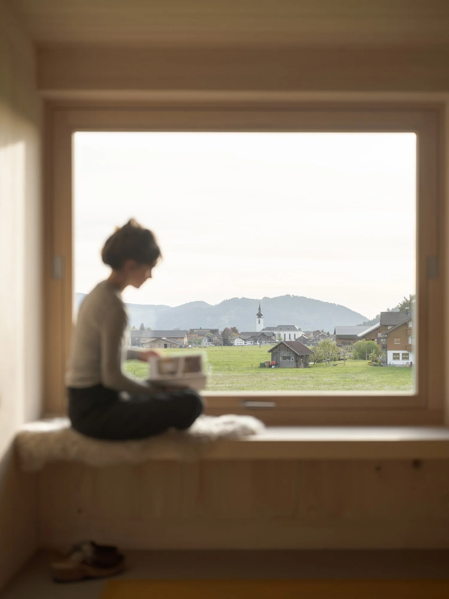 Blick durch ein Fenster auf eine ländliche Landschaft mit Häusern, Wiesen, Bäumen, Bergen im Hintergrund und einem Kirchturm in der Mitte.