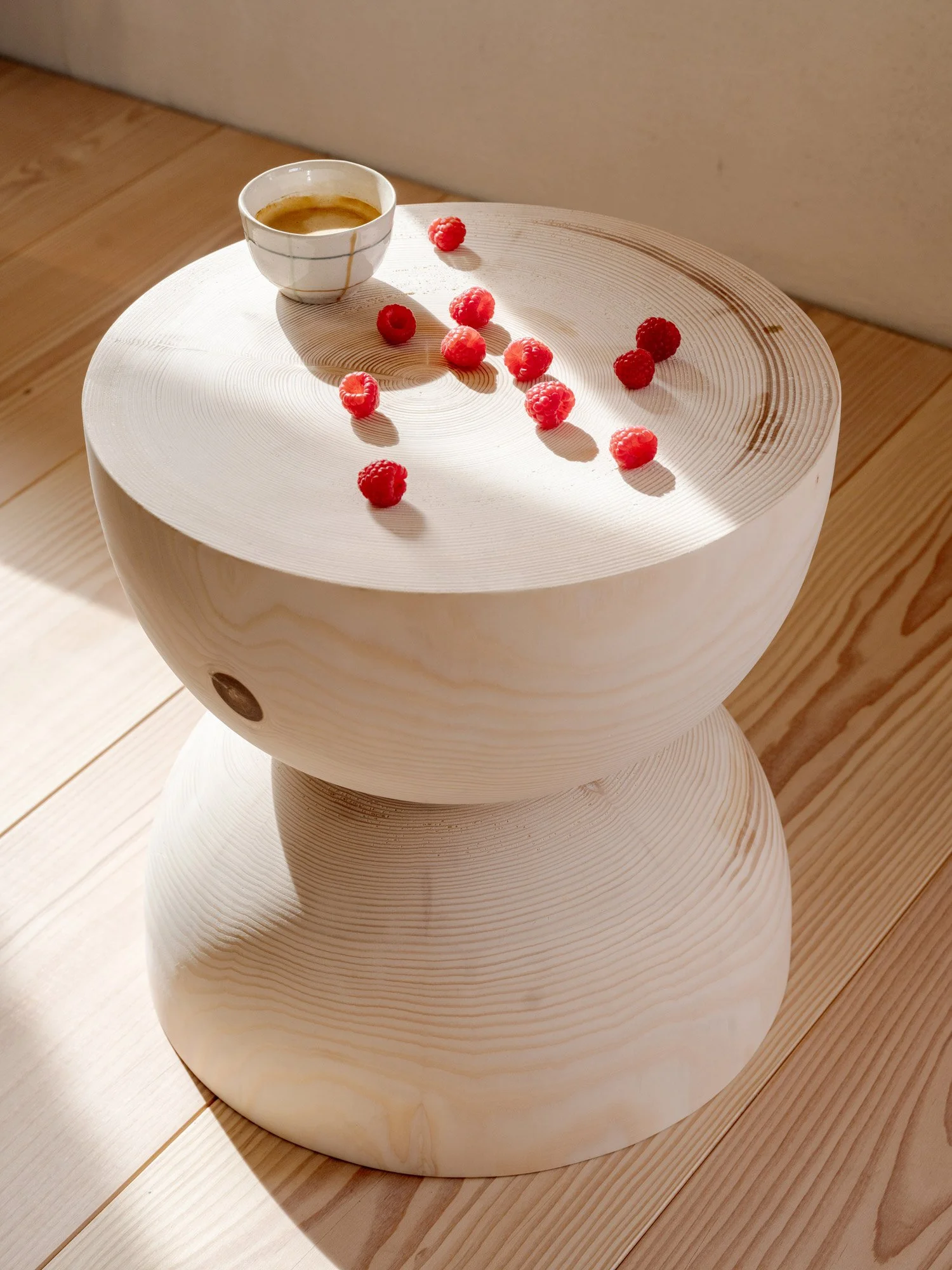 Eine weiße, runde Holzskulptur mit einer flachen Oberfläche, auf der mehrere Himbeeren verteilt sind. Im Hintergrund steht eine Tasse Kaffee, die leicht angedunkelt ist. Die Szene ist von natürlichem Licht beleuchtet.