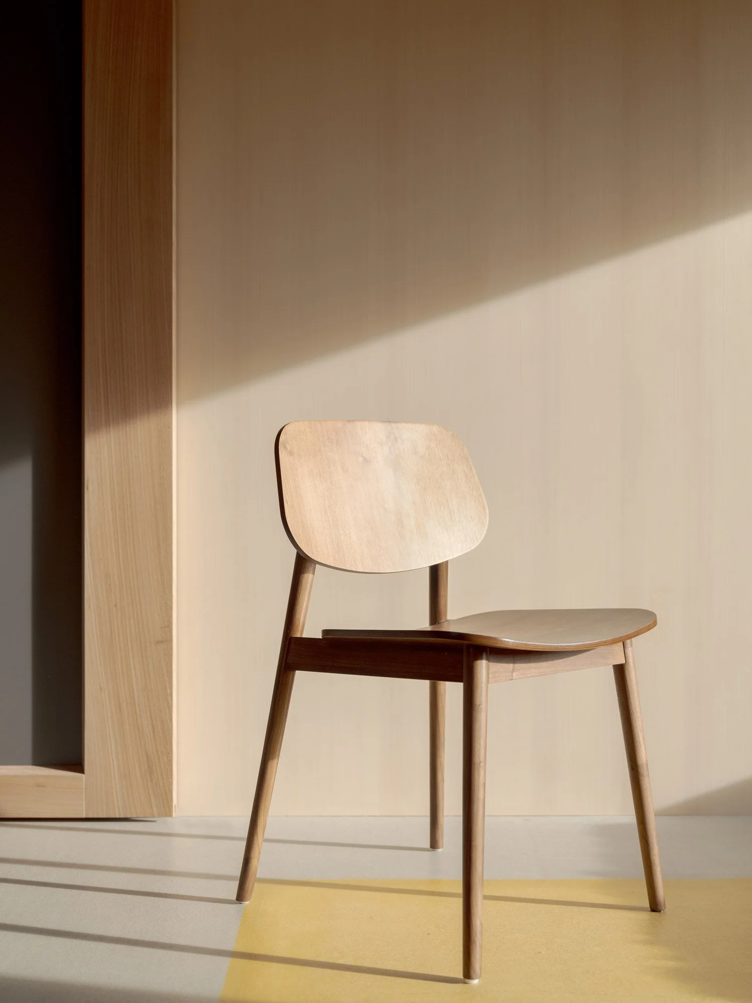 Ein Stuhl aus Holz mit schräg gestellten Beinen vor einer hellen Wand, Licht und Schatten fallen auf den Stuhl und die Wand.