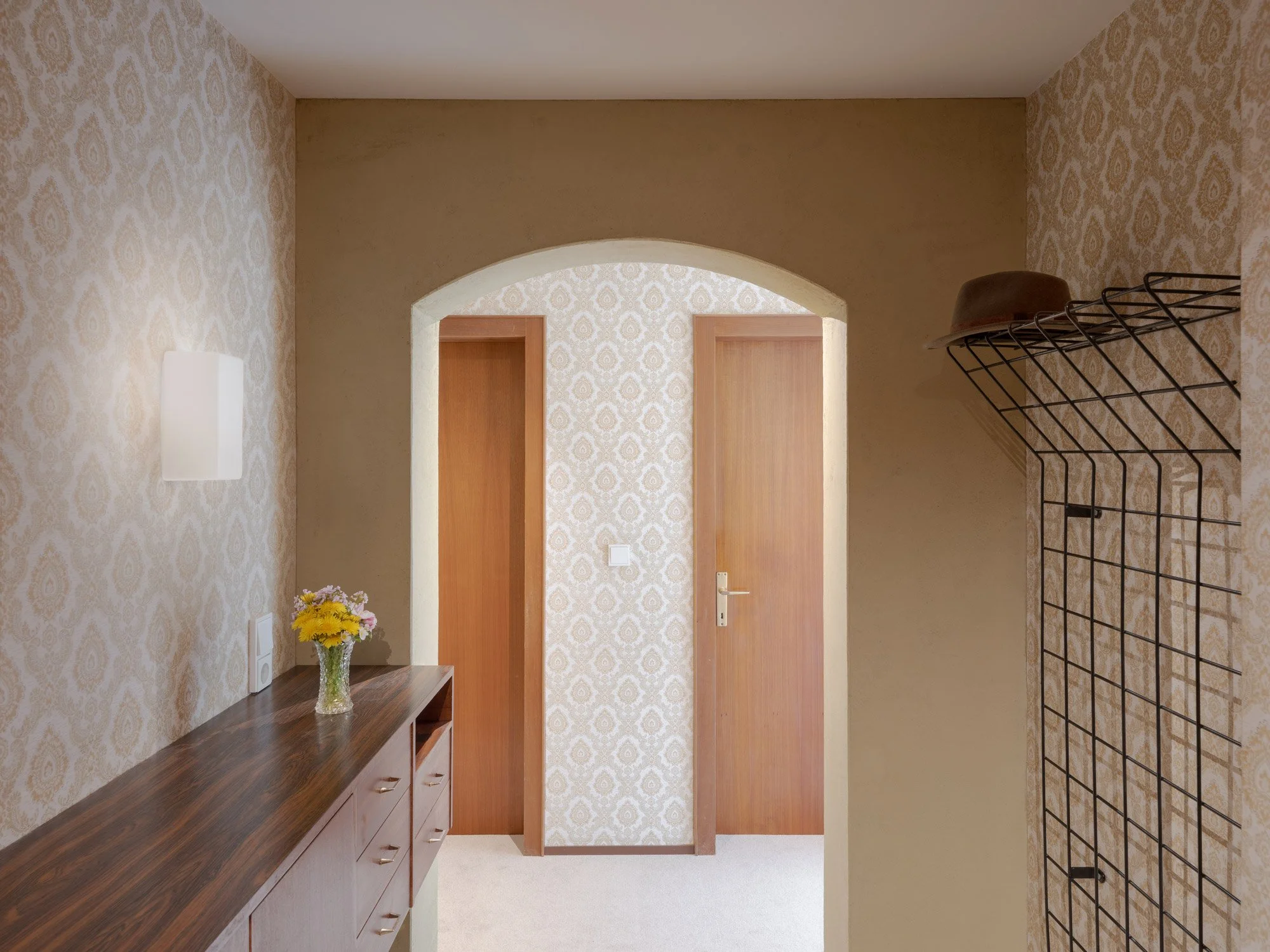 Ein Flur mit gemusterten Tapeten, Holztüren, einem Holzsideboard mit Blumenvase und eine Wandleuchte. Zur rechten Seite ein Möbeltisch mit Metalldrahtregal.