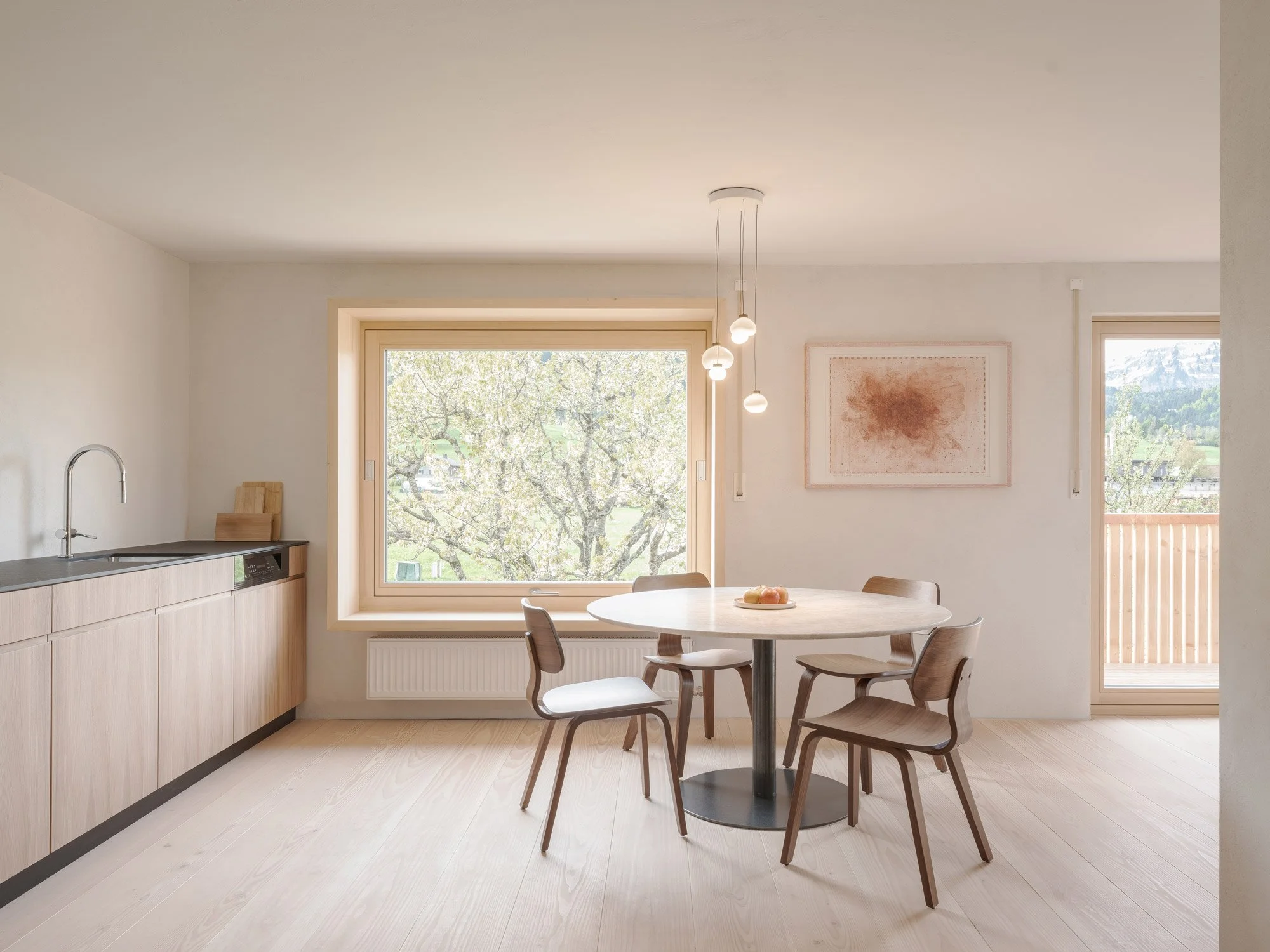 Innenaufnahme einer modernen, hellen Küche mit Essbereich, Holzofen, runden Holztisch mit vier Stühlen, großen Fenstern mit Blick auf Bäume und Berge, zeitgenössische Kunst an der Wand und Deckenleuchte.