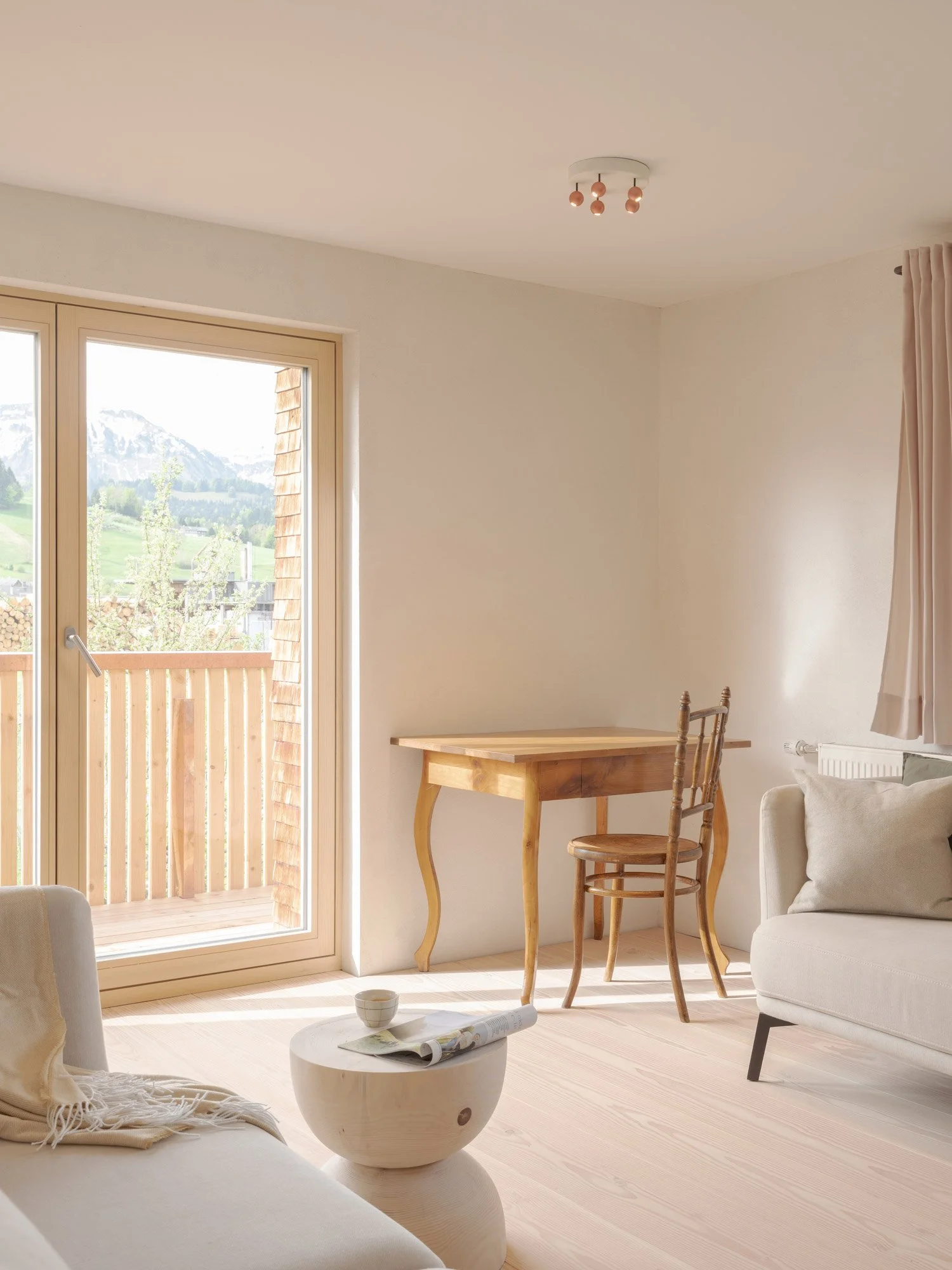 Helles Wohnzimmer mit großen Fenstern, Holztisch, Stühlen, Sofa und einem Couchtisch mit einer Schale und Zeitung. Blick auf die Berge im Hintergrund.