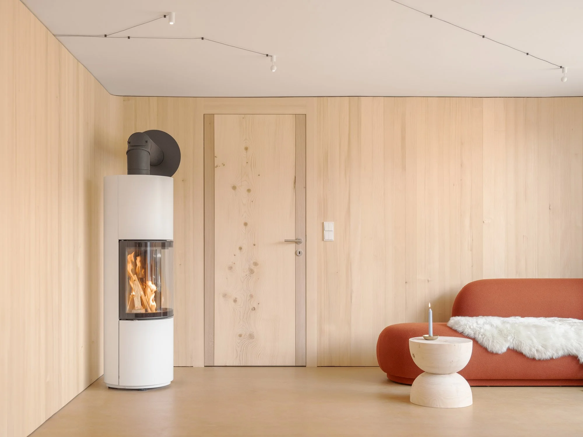 Ein modernes Wohnzimmer mit einem Holzfussboden, einer roten Couch, einem weißen Teppich, einem kleinen weißen Tisch und einem Kamin, der Feuer speichert. Die Wände sind aus hellem Holzplatten, die Tür ist ebenfalls aus Holz. An der Decke hängen dünne Kabel mit kleinen weißen Leuchten.