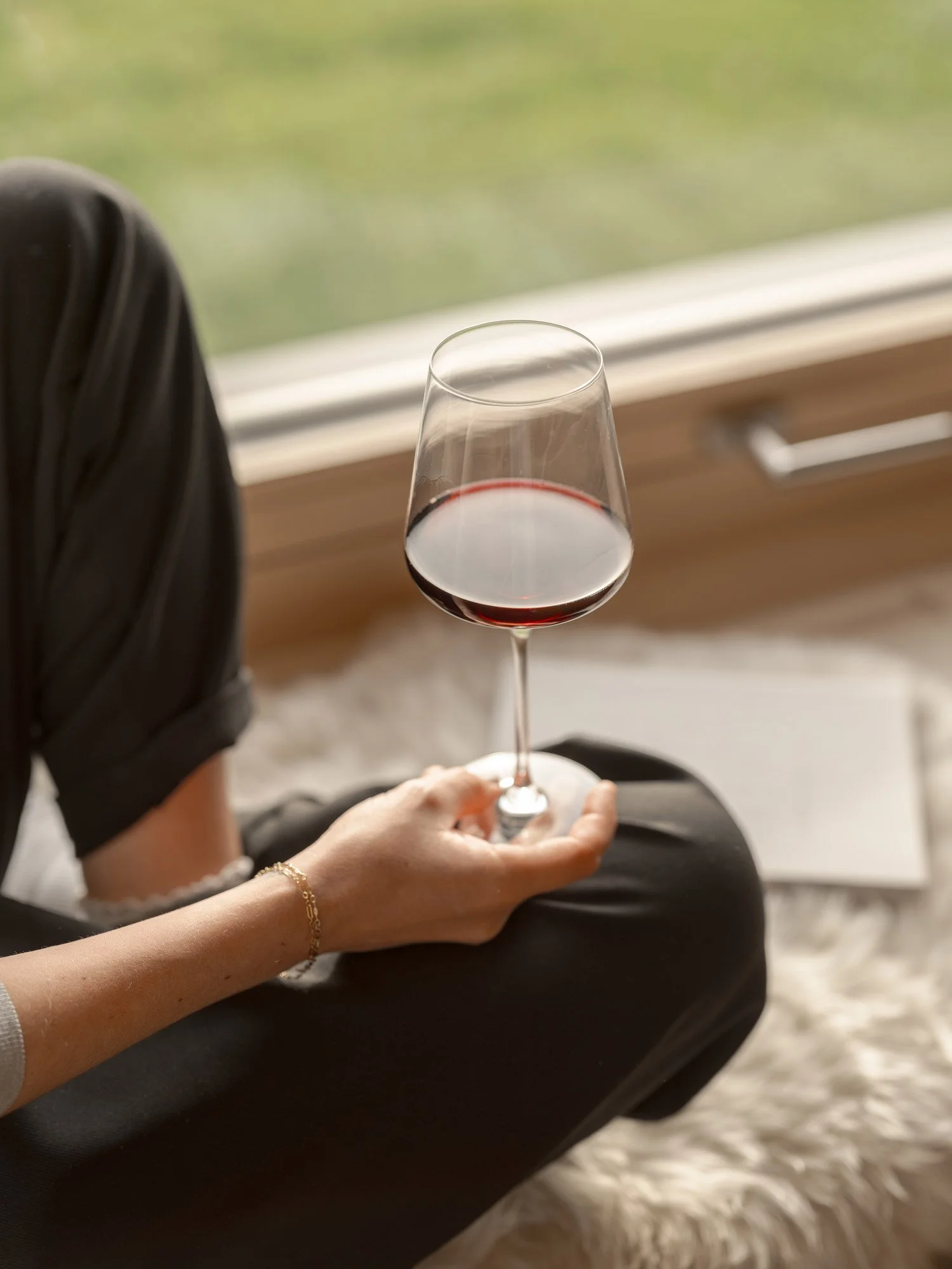 Eine Person hält ein Weinglas mit Rotwein, während sie auf einem Stuhl sitzt, neben einem Fenster mit Blick nach draußen, im Hintergrund ein grüner Rasen.