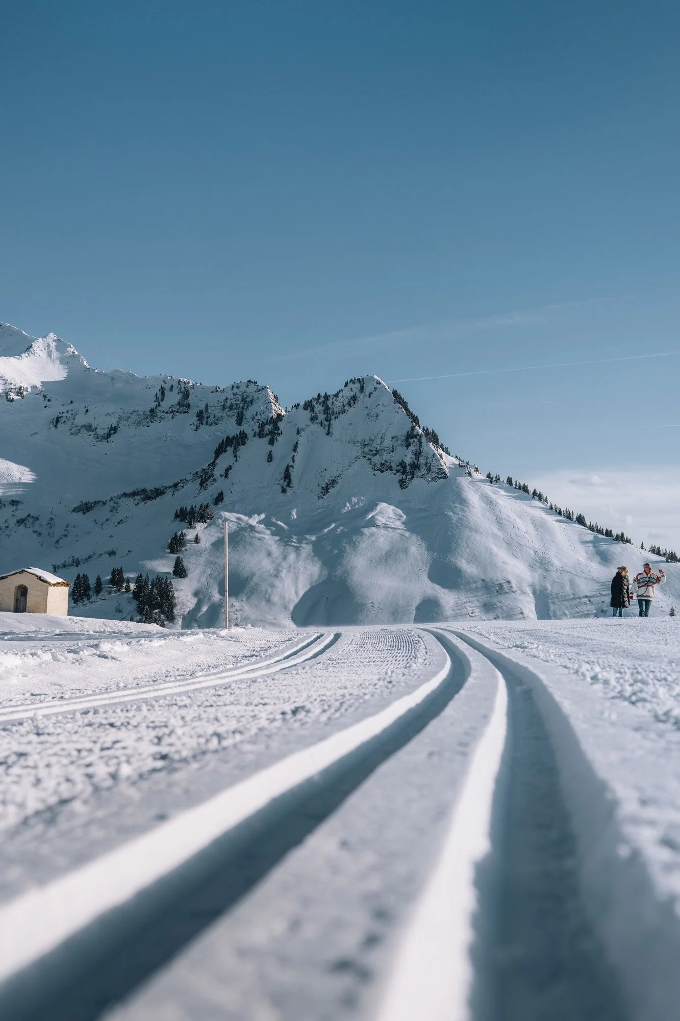 Schneebedeckte Berge mit klarer blauer Himmel, Skispuren im Schnee, kleiner Haus und zwei Personen im Wintersportoutfit im Vordergrund.