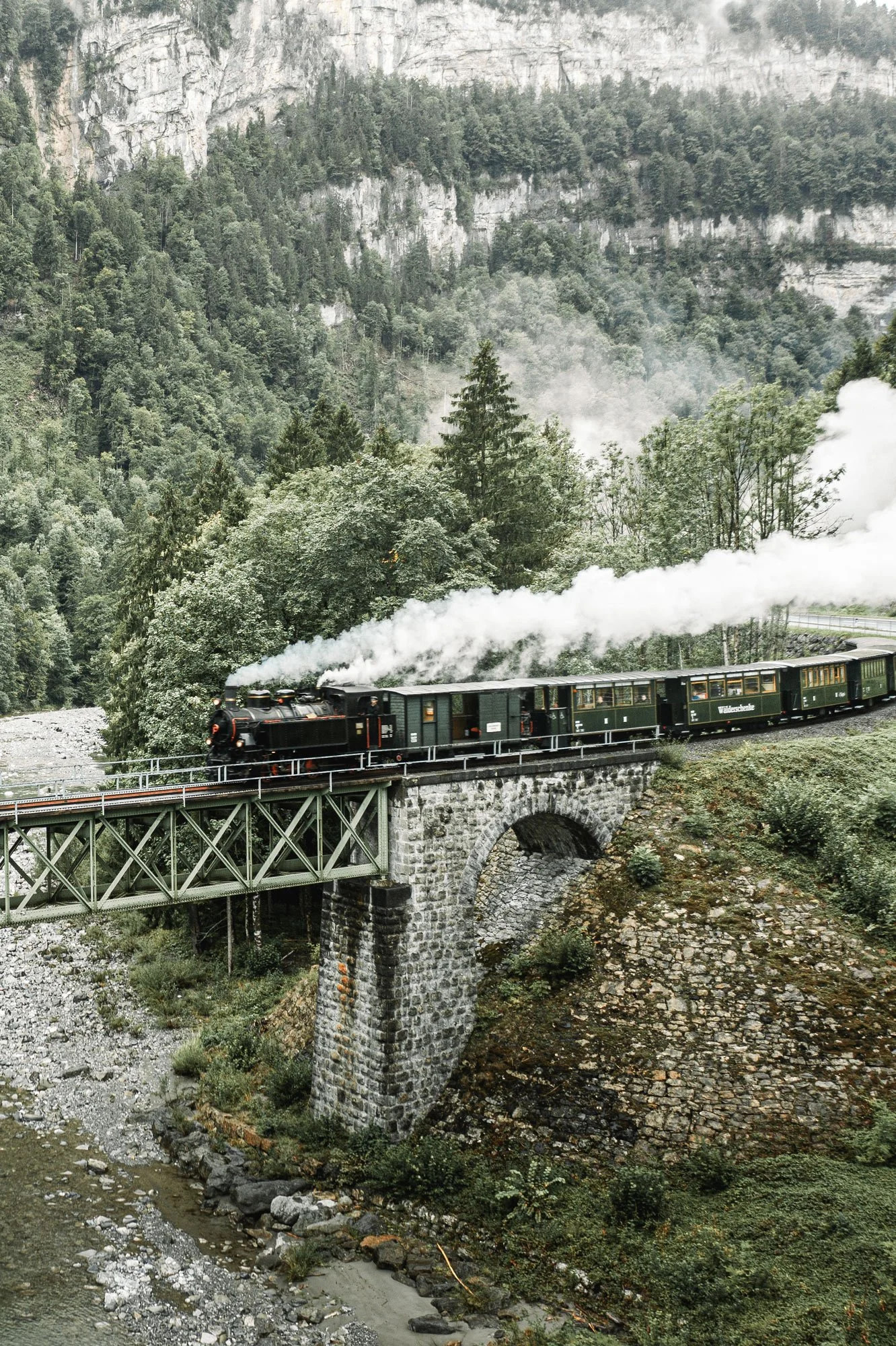 Ein Dampflokomotive fährt über eine steinerne Brücke in einer bergigen, bewaldeten Landschaft, mit Fluss unterhalb der Brücke und Nebel in der Luft.