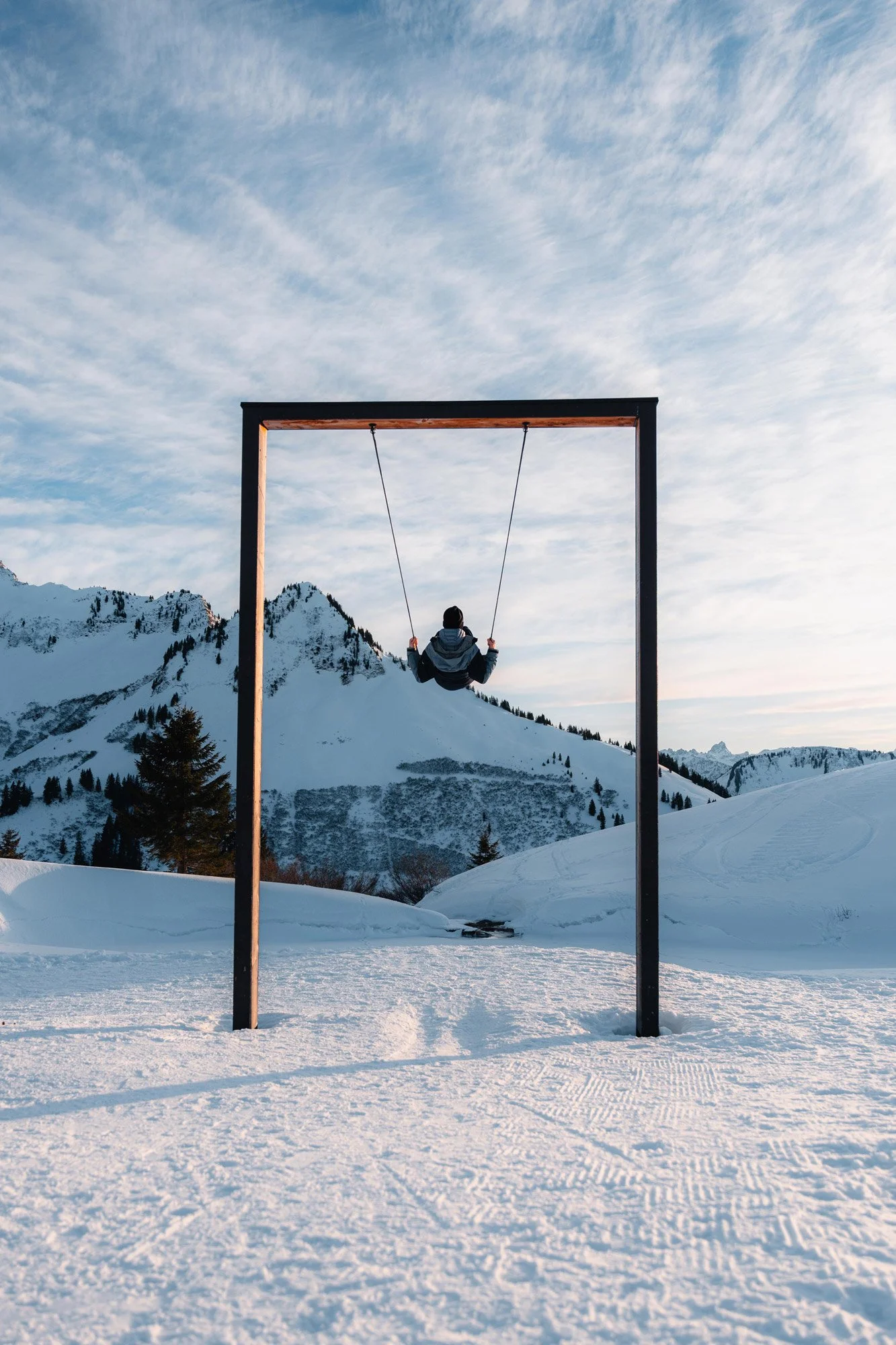 Ein Kind schaukelt in einem Skigebiet mit schneebedeckten Bergen im Hintergrund, während die Sonne untergeht oder aufgeht, im Winter.