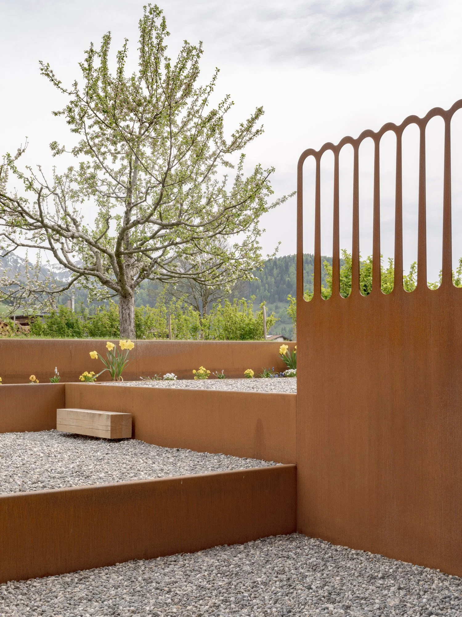 Ein Garten mit einem blühenden Baum, einer hohen rustikalen Metallzaun, Blumenbeeten und einem Kiesweg, im Hintergrund grüne Hügel und ein bewölkter Himmel.