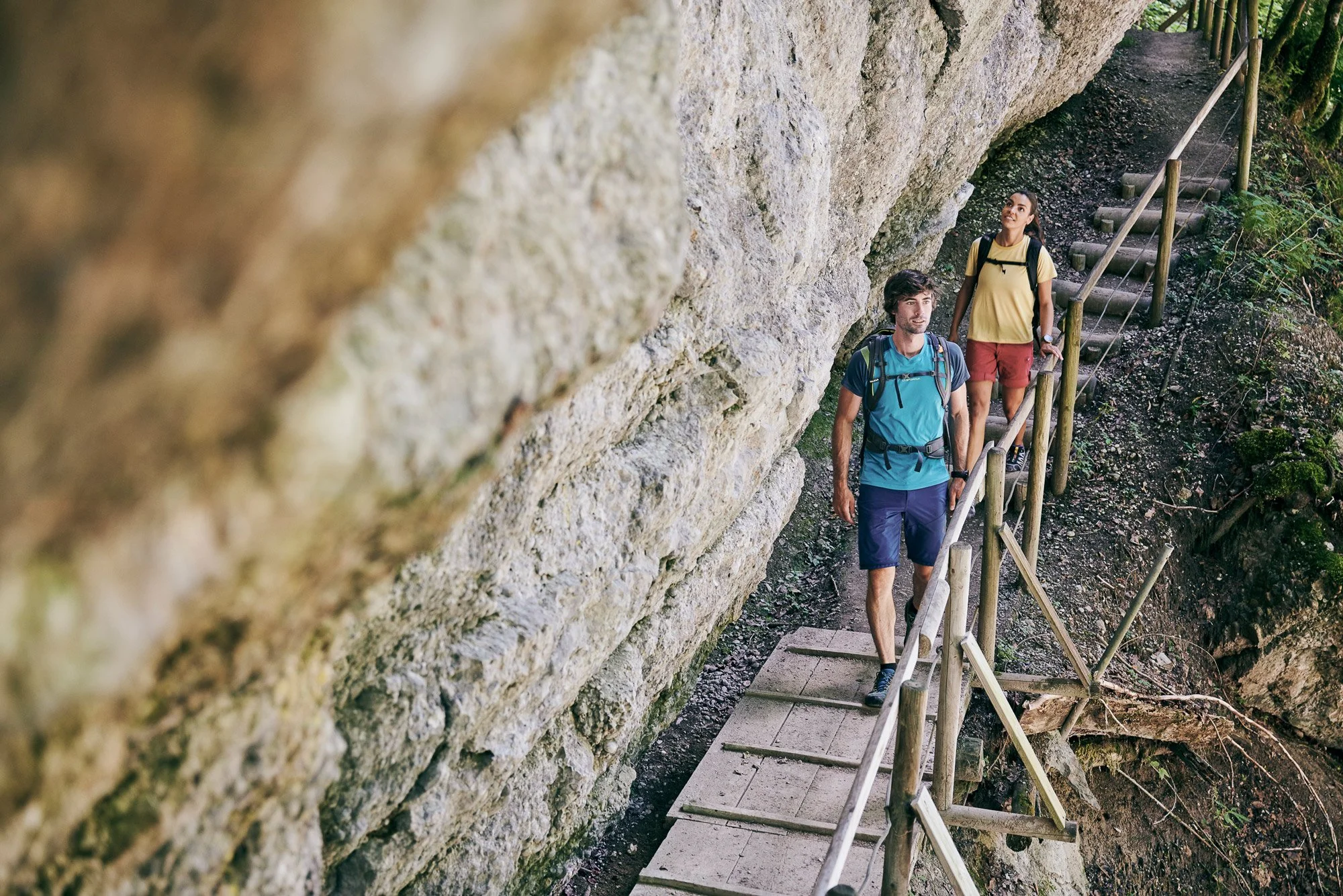 Zwei junge Männer wandern auf einem steilen Weg entlang einer Felswand, gesichert mit einem Geländer, in einem bewaldeten Gebiet.