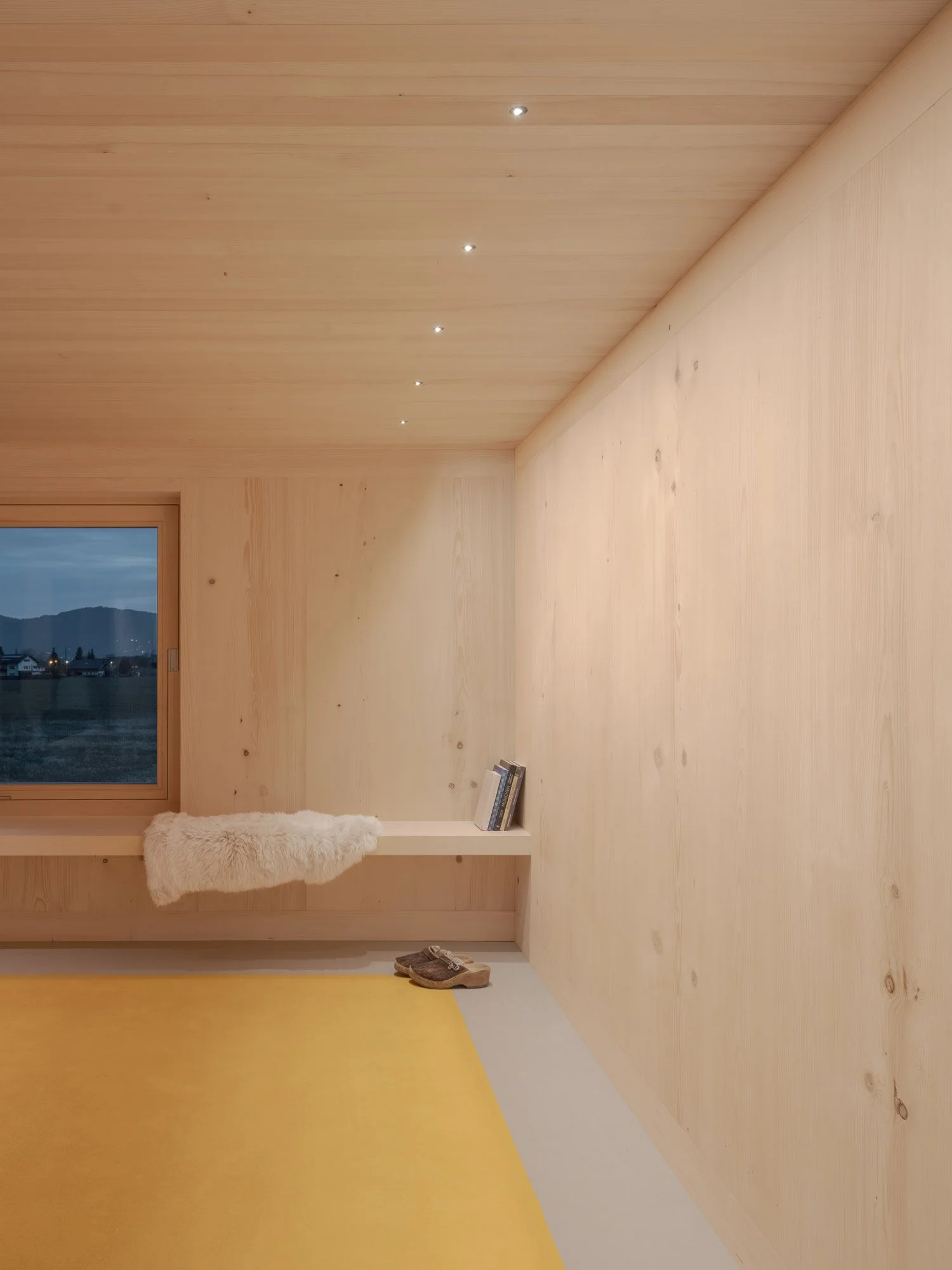 Innenraum mit Holzverkleidung, ein Fenster mit Bergblick, ein Regal mit Büchern, ein Fellteppich, eine gelbe und weiße Bodenfläche, Schuhe auf dem Boden.