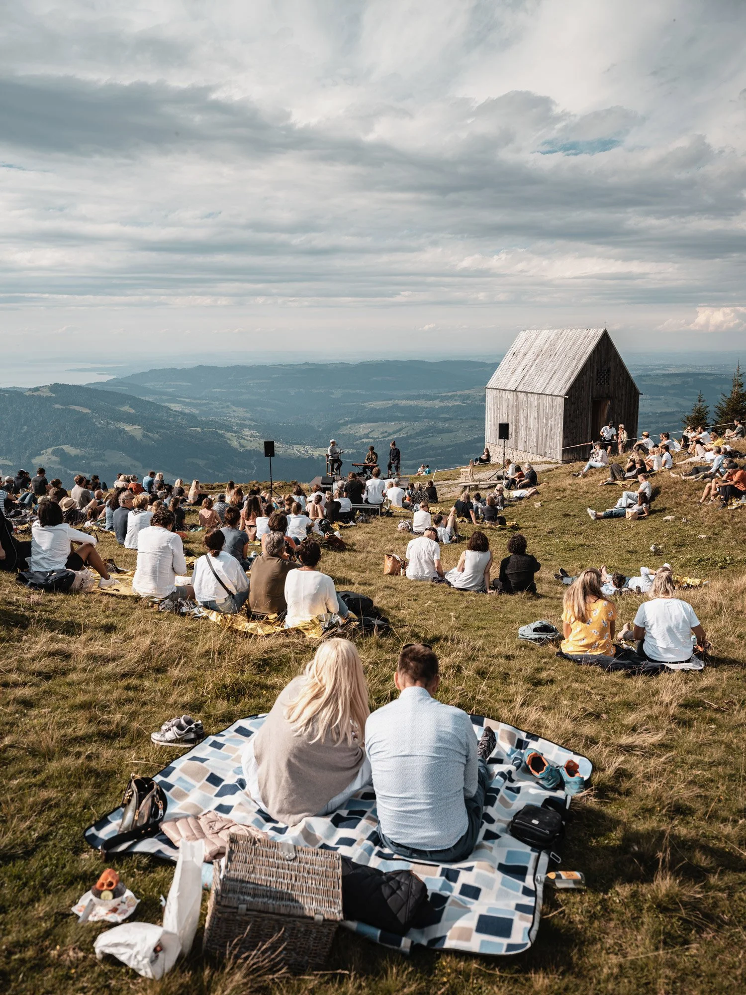 Viele Menschen sitzen auf einer Wiese im Freien und schauen zu einer kleinen Bühne, auf der jemand Musik macht. Es ist eine Szene in einer bergigen Landschaft mit Wolken am Himmel.