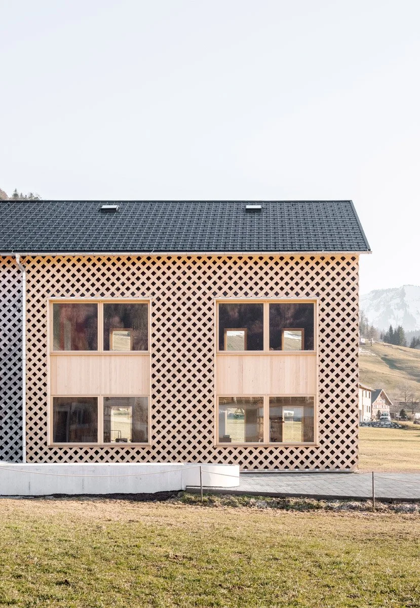 Ein modernes Wohnhaus mit Holzverkleidung und einem Gittermuster an der Außenfassade, große Fenster, eine dunkle Dachziegeldeckung, umgeben von einem Rasen und einem ländlichen Umfeld.