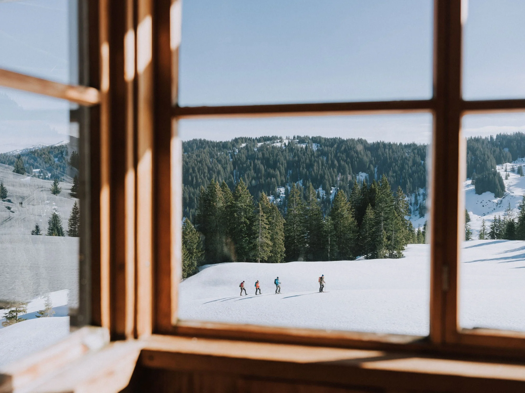 Blick durch ein Fenster auf eine verschneite Berglandschaft mit Wäldern und Skifahrern im Vordergrund.