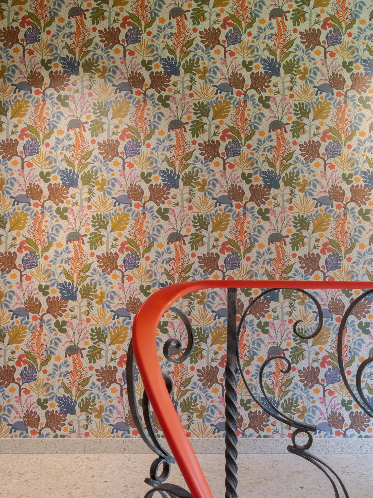 Blick auf eine Wand mit buntem, floralen Tapetenmuster und einen roten, spiralförmigen Handlauf im Vordergrund.