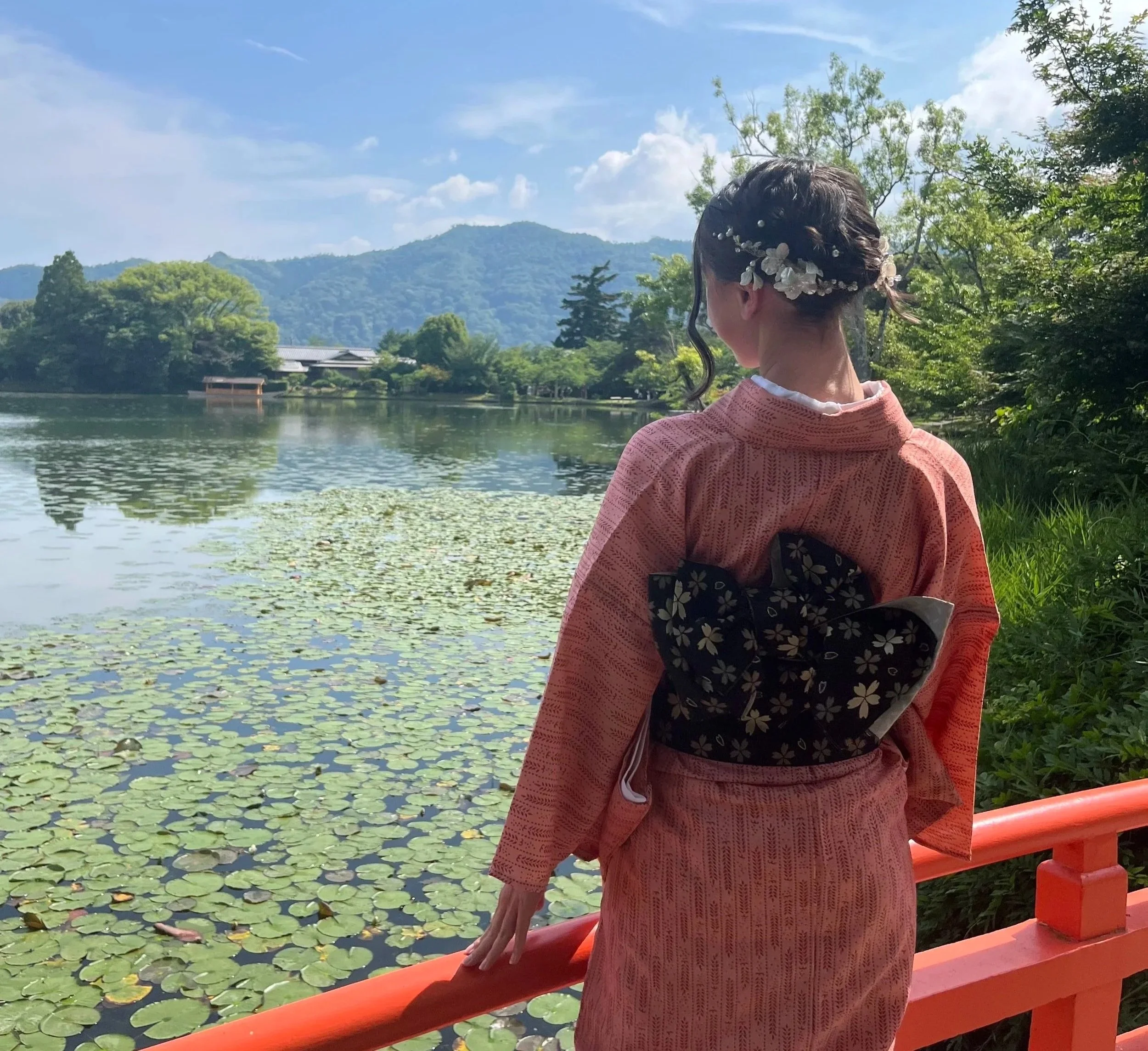 A woman dressed in traditional Japanese clothing, with floral hair accessories, stands near a lake with lotus leaves, looking out at mountains and a partly cloudy sky.