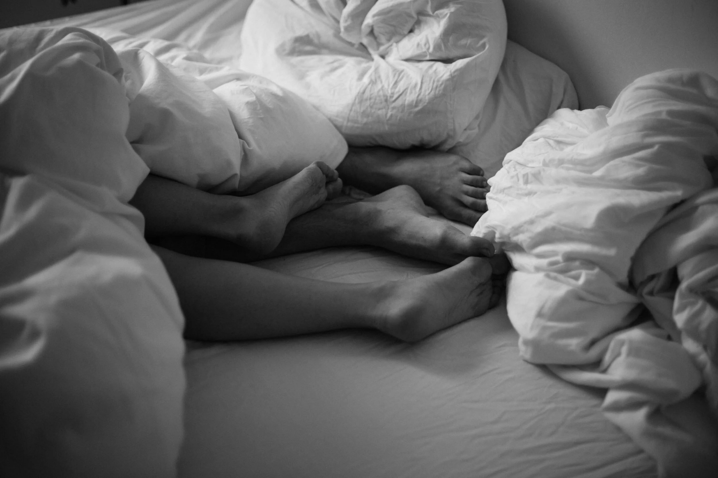 Disorderly bed with crumpled white sheets and bedding, partially showing a person's hand and foot beneath the sheets. Sex underneath the sheets.