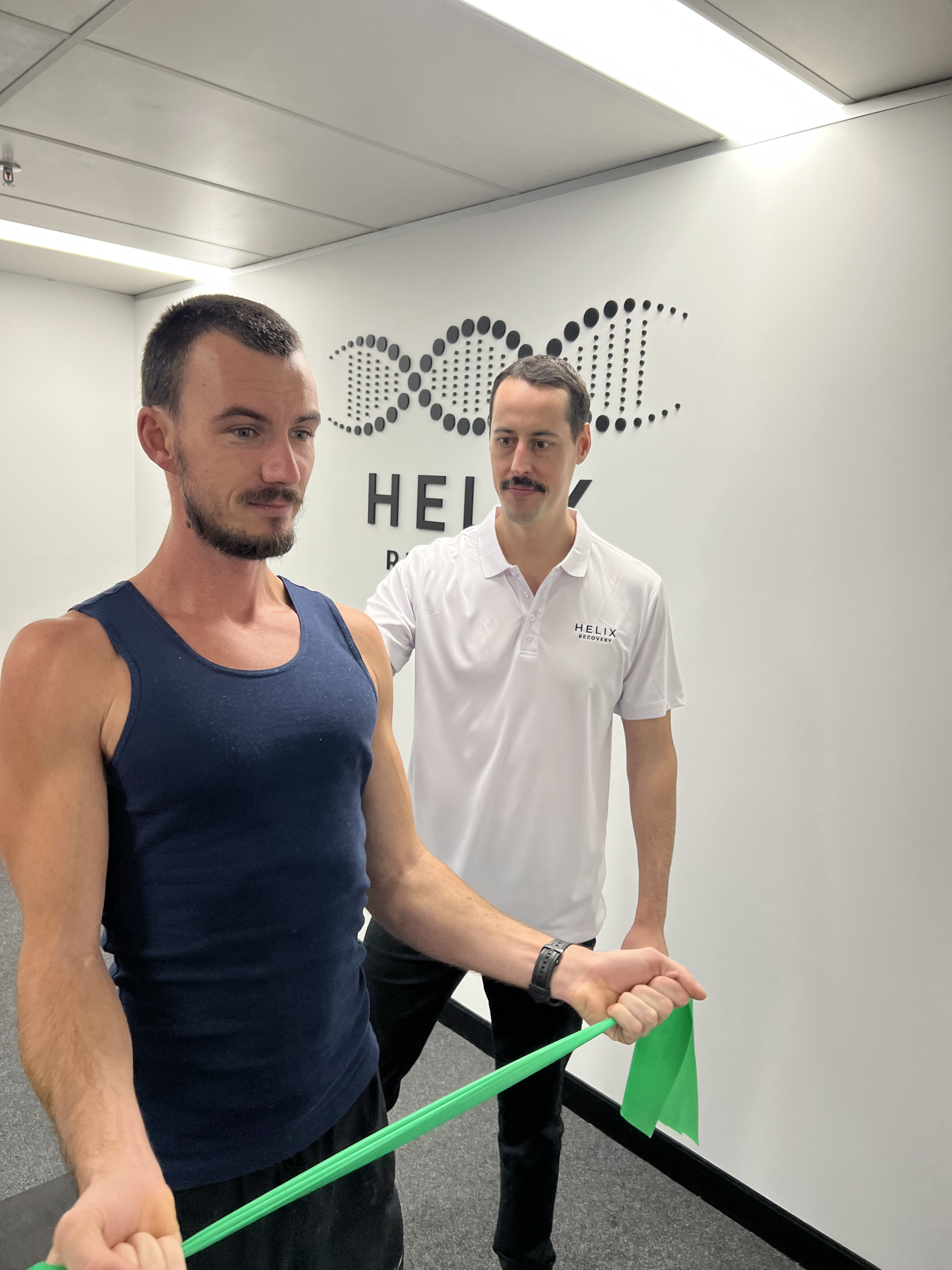 A practitioner shows an exercise to a patient with a HELIX Recovery sign on the wall behind them. One man is wearing a dark tank top, and the other is wearing a white shirt with the HELIX Recovery logo.