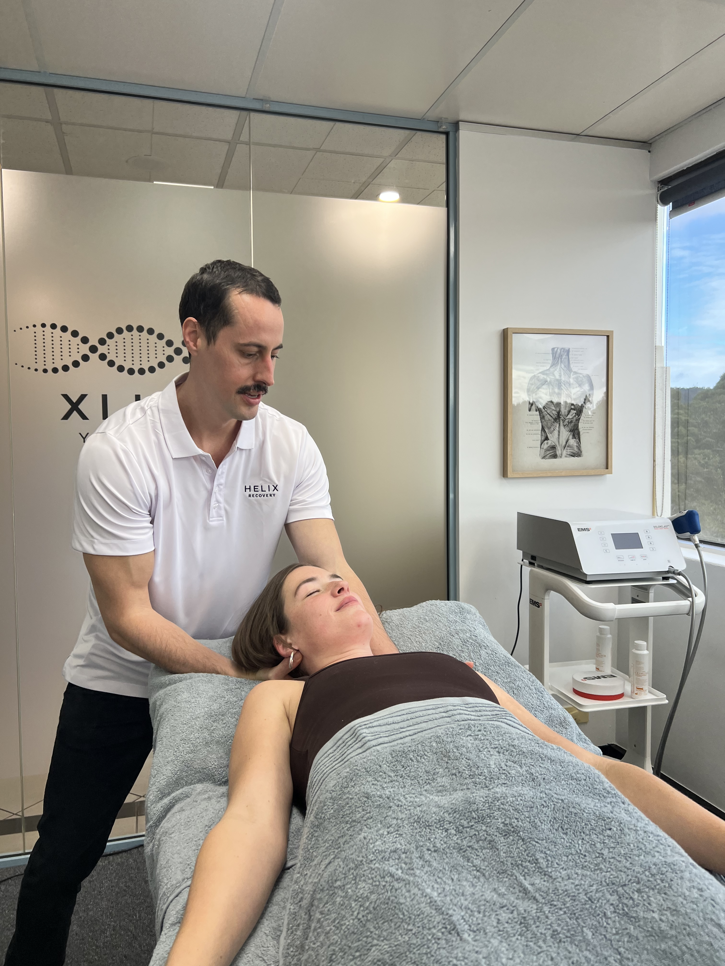 A medical professional is performing a treatment or exam on a woman lying on a bed, in a clinical setting with medical equipment and a framed anatomical chart on the wall.