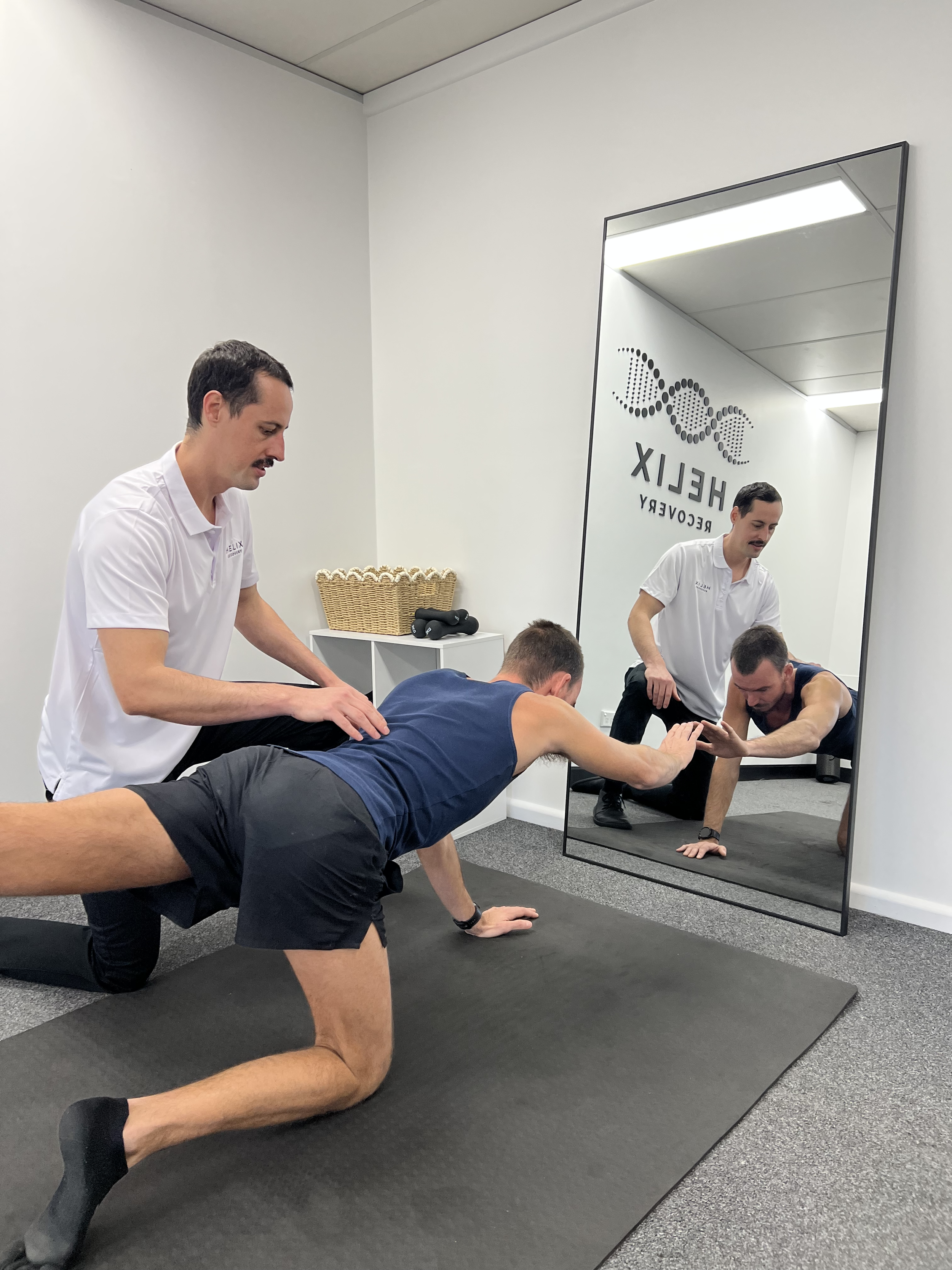 Two men are doing a plank exercise on black yoga mats on the floor. An instructor in a white shirt is assisting them, and their image is reflected in a large mirror on the wall. The room has white walls, and there's a shelf with a basket and some black objects.