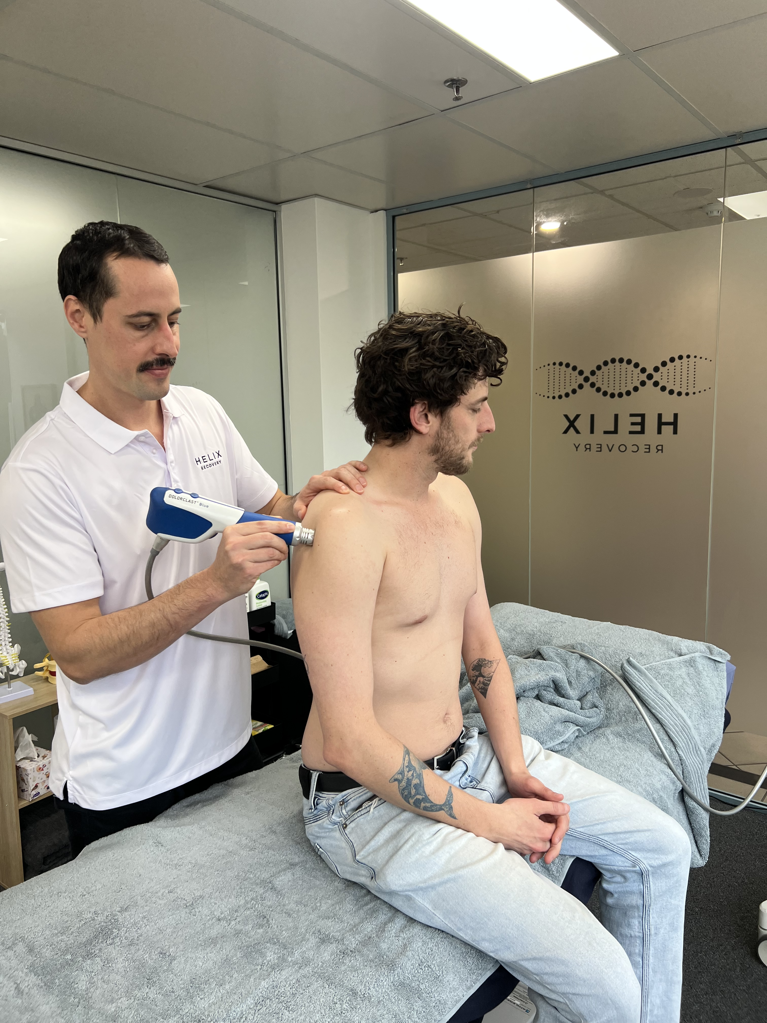 A man sits shirtless on an examination table at a medical clinic, while a healthcare professional in a white shirt prepares to perform a procedure on his upper back with a handheld device. The clinic has a logo for Helix Recovery on the glass wall.