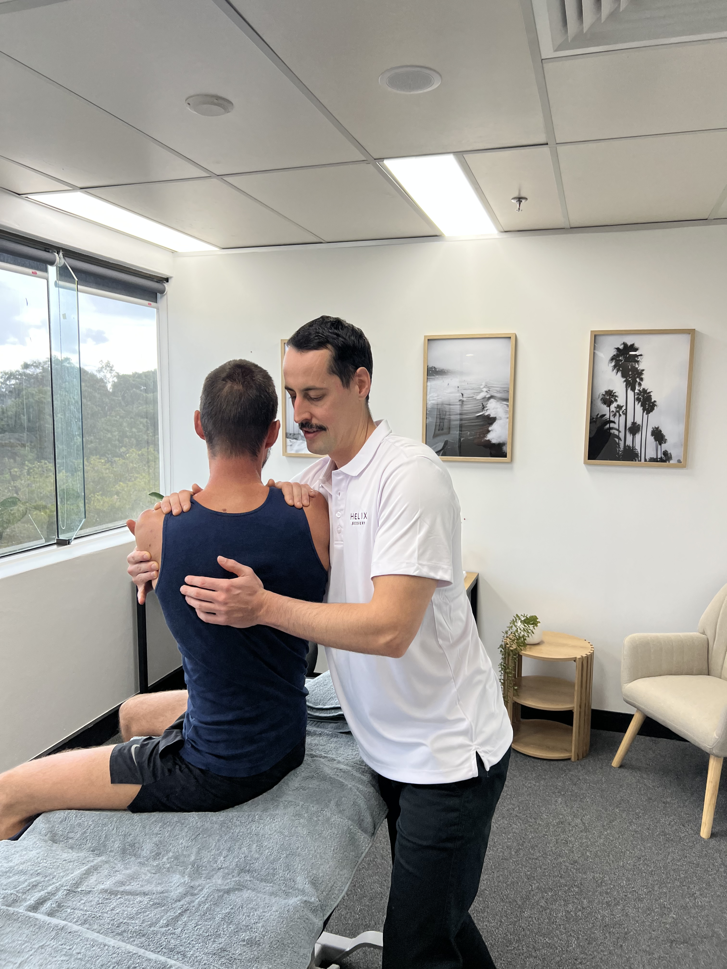 An osteopath adjusting a patient's back in an office with framed pictures of beaches and palm trees on the wall.