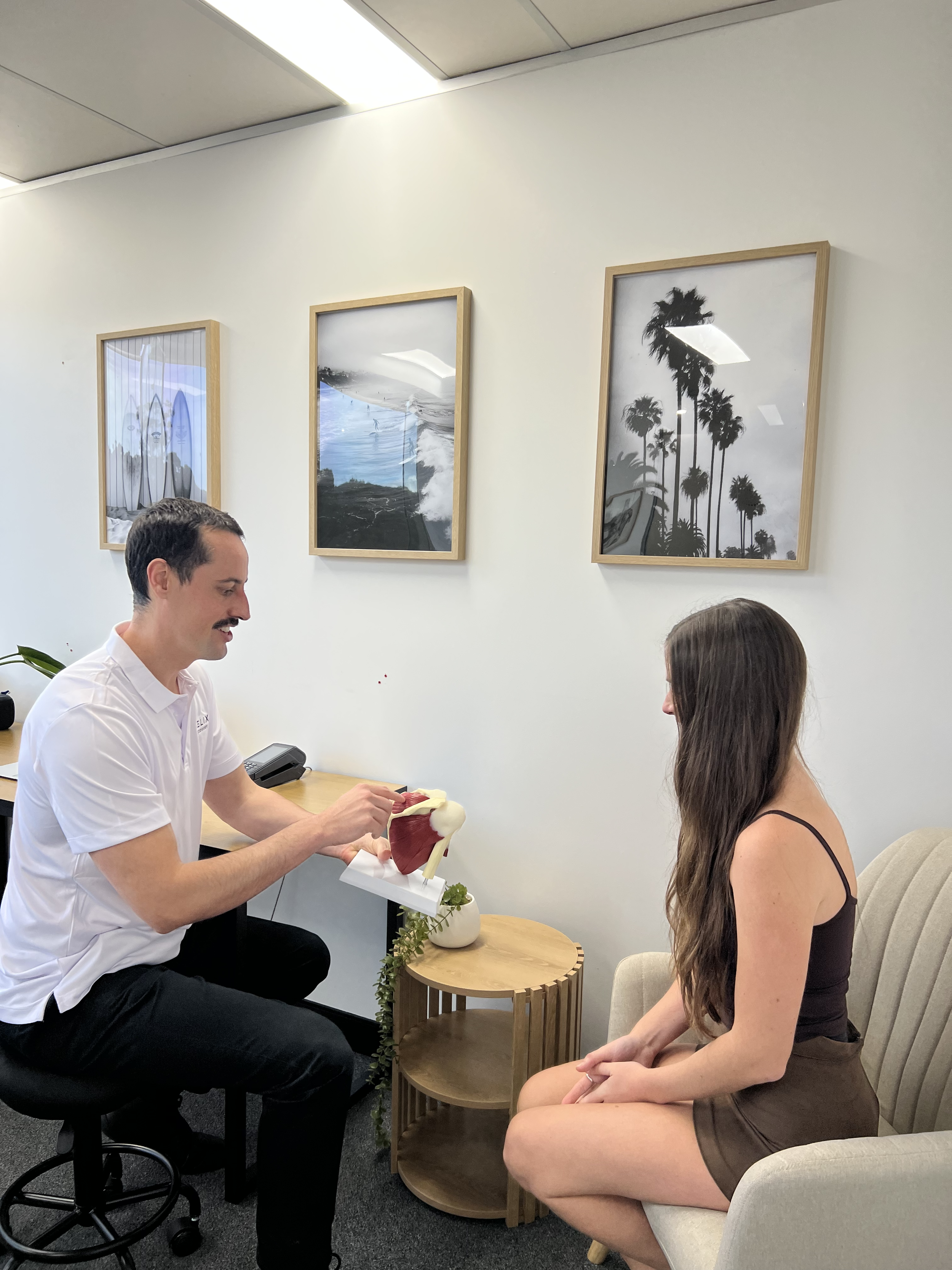 A man in a white shirt and black pants is showing a model to a woman in a brown dress. They are seated in a room with framed black-and-white photographs of palm trees and a beach scene on the wall behind them, with a small round wooden side table between them.