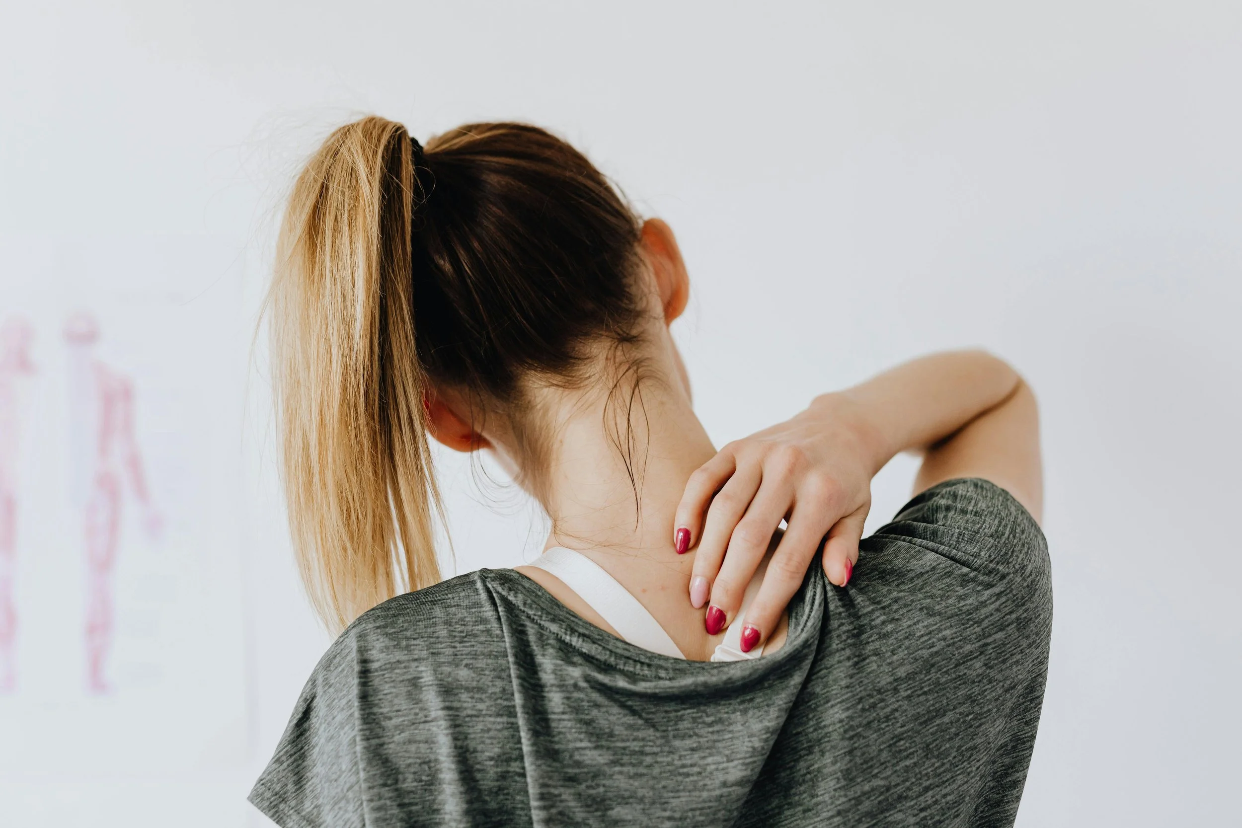 A woman with her hair tied back in a ponytail, wearing a gray top, is stretching or rubbing her neck with one hand, seen from behind against a plain white wall.