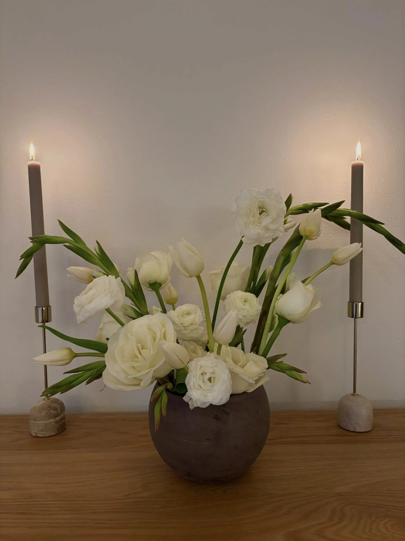A floral arrangement of white flowers, including lilies and lisianthus, in a dark ceramic vase on a wooden surface, flanked by two tall candles in marble holders.