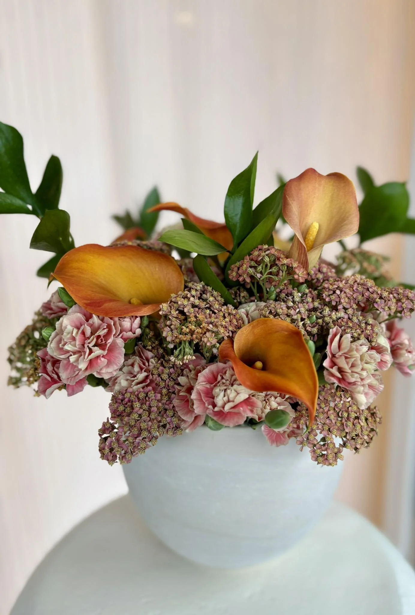 A white vase holding a bouquet of pink carnations, orange calla lilies, and green leaves against a light background.