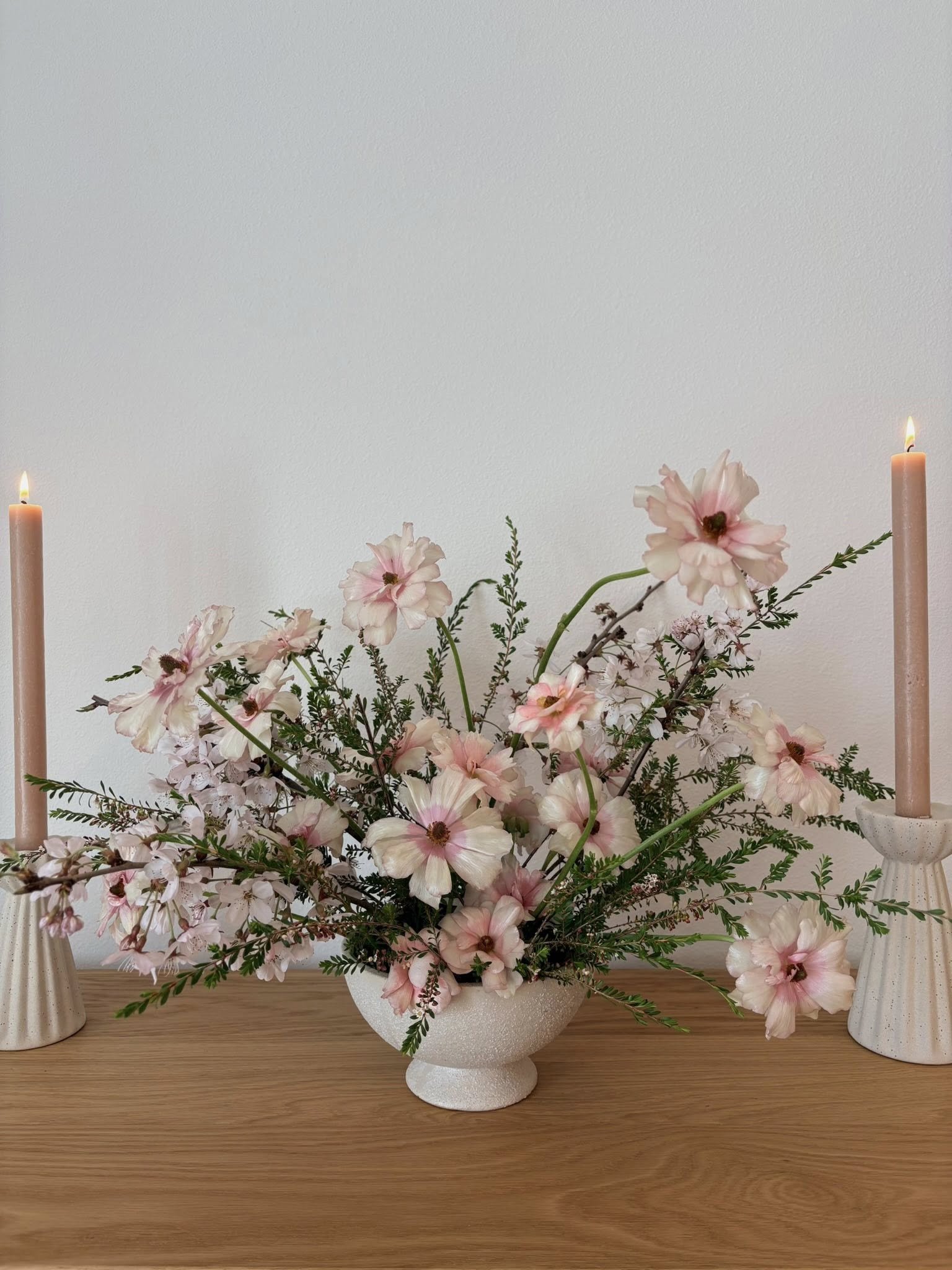 A floral arrangement of pink and white flowers with green foliage in a white textured vase, flanked by two tall pink candles on white candle holders on a wooden surface.