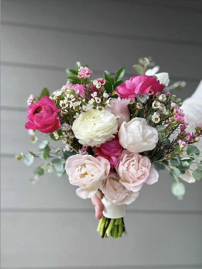 A bouquet of pink, white, and cream flowers with green foliage, held by a person in front of a gray background.