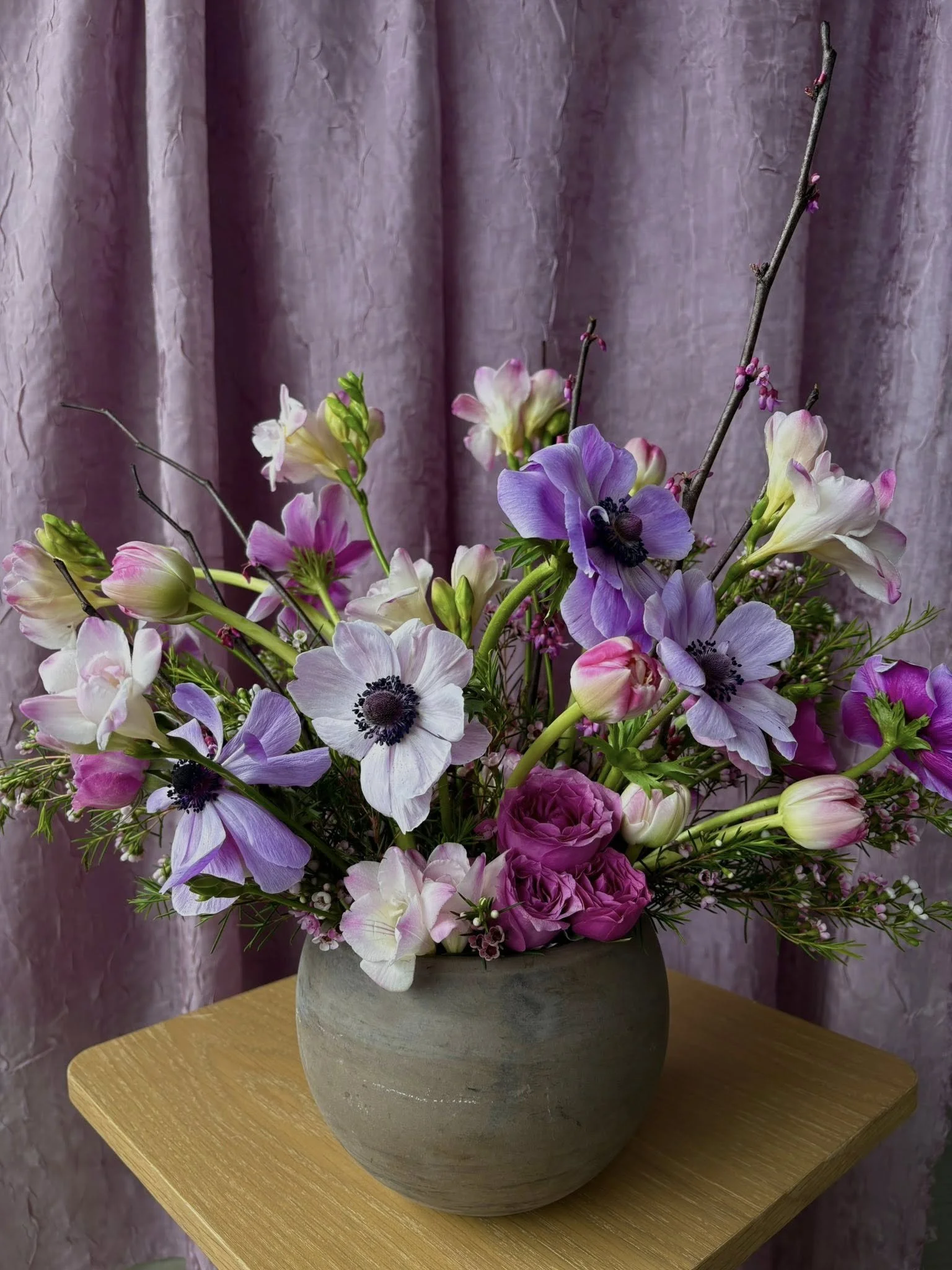 A large bouquet of pink, purple, white, and green flowers in a rounded gray vase on a wooden table with a pink curtain in the background.