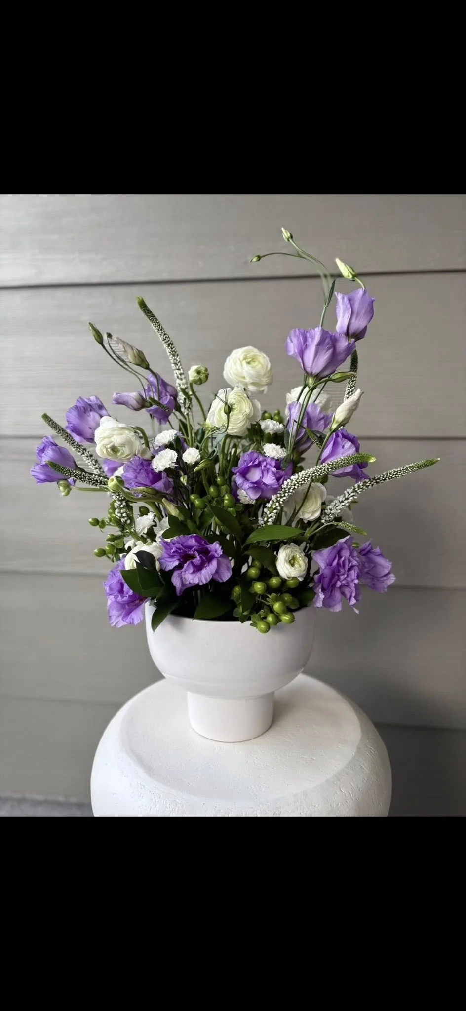 A white ceramic vase filled with a bouquet of purple and white flowers, featuring lisianthus, greenery, and small berries, placed on a white pedestal against a gray wooden background.
