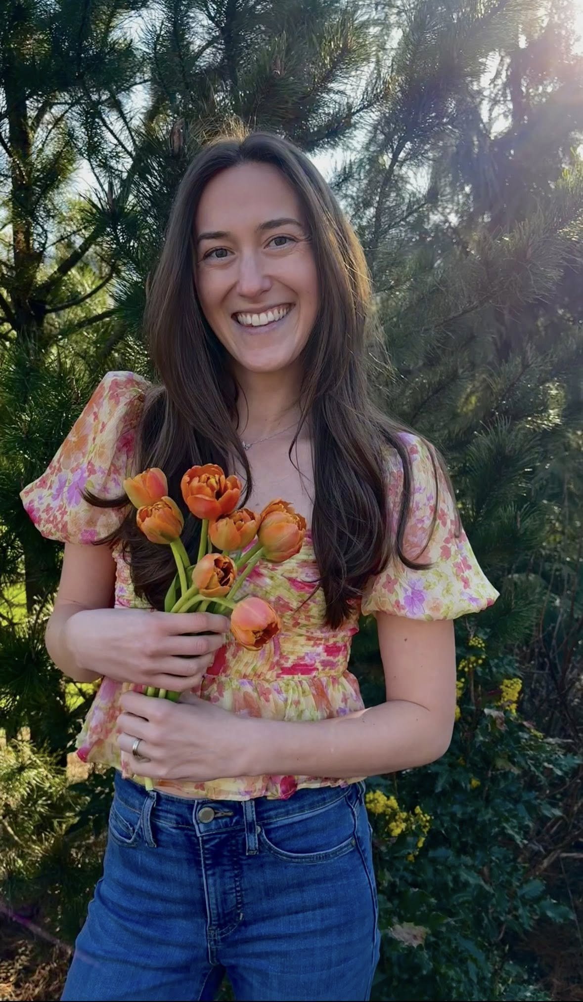 A young woman with long brown hair smiling, holding a bunch of orange tulips, wearing a yellow floral top and blue jeans, standing outdoors in front of a pine tree with sunlight filtering through the branches.