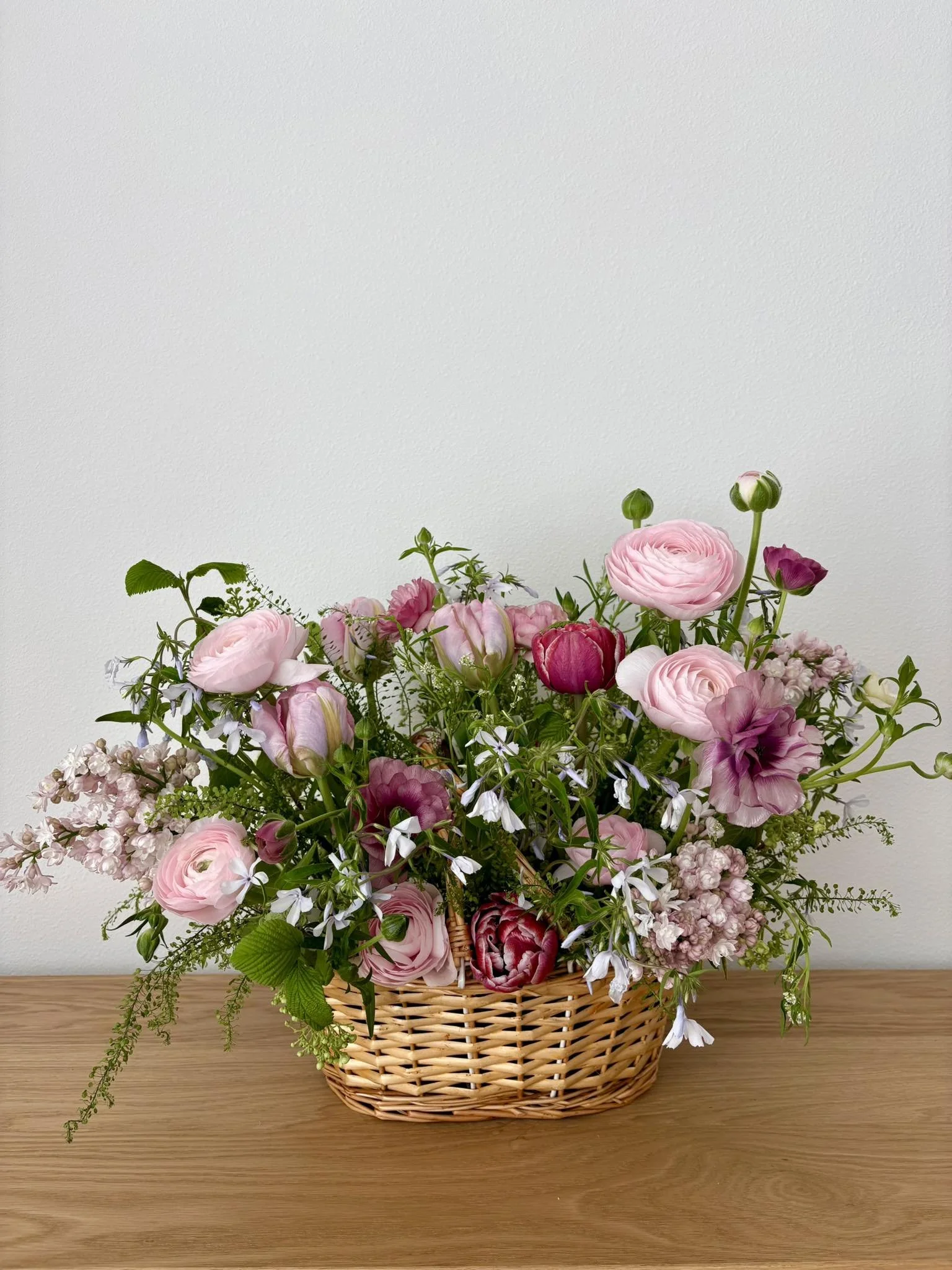 A basket filled with pink and purple flowers, sitting on a wooden surface against a plain white wall.