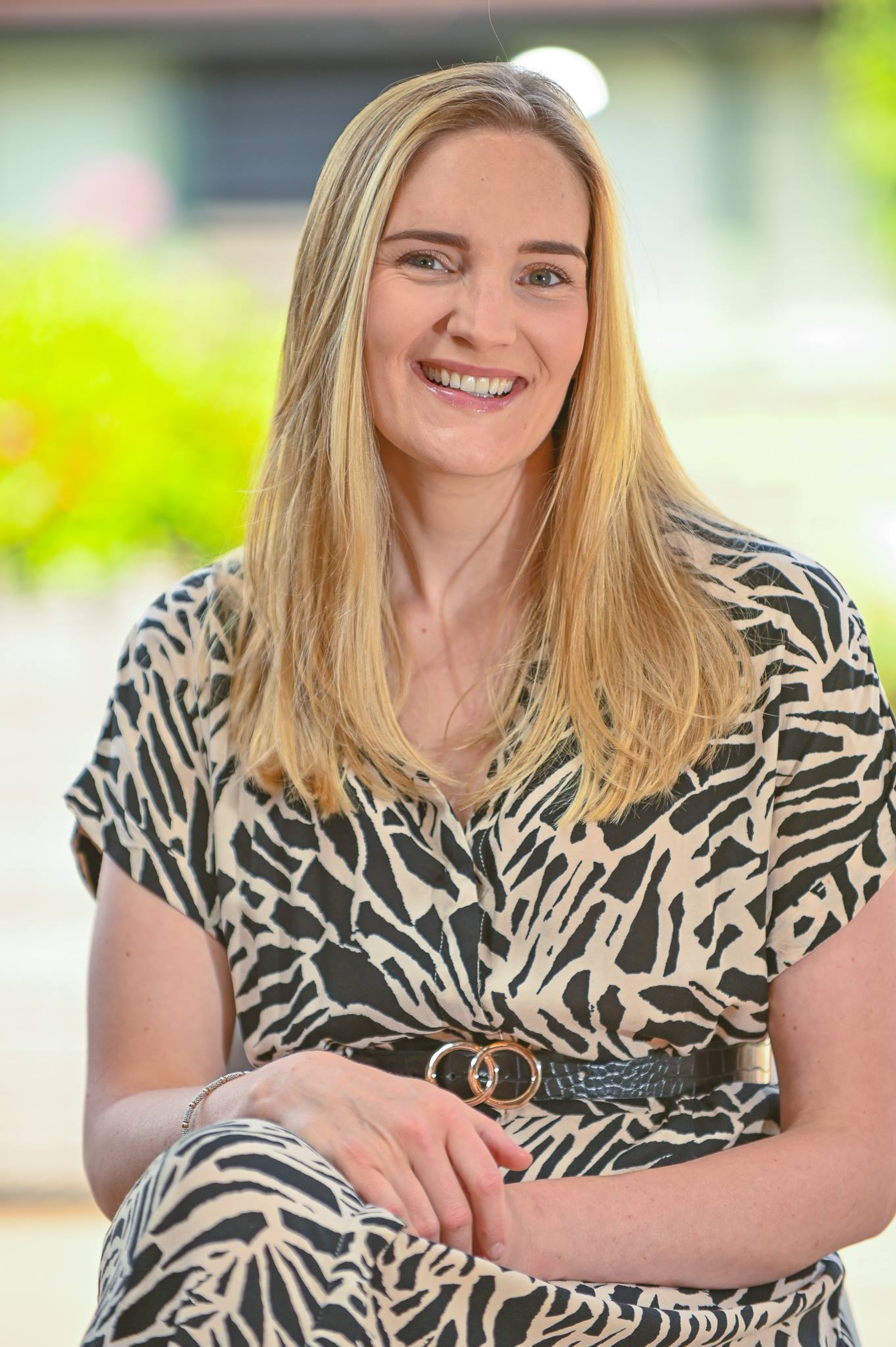 A smiling woman with long blonde hair wearing a black and white animal print dress, sitting outdoors during daytime.