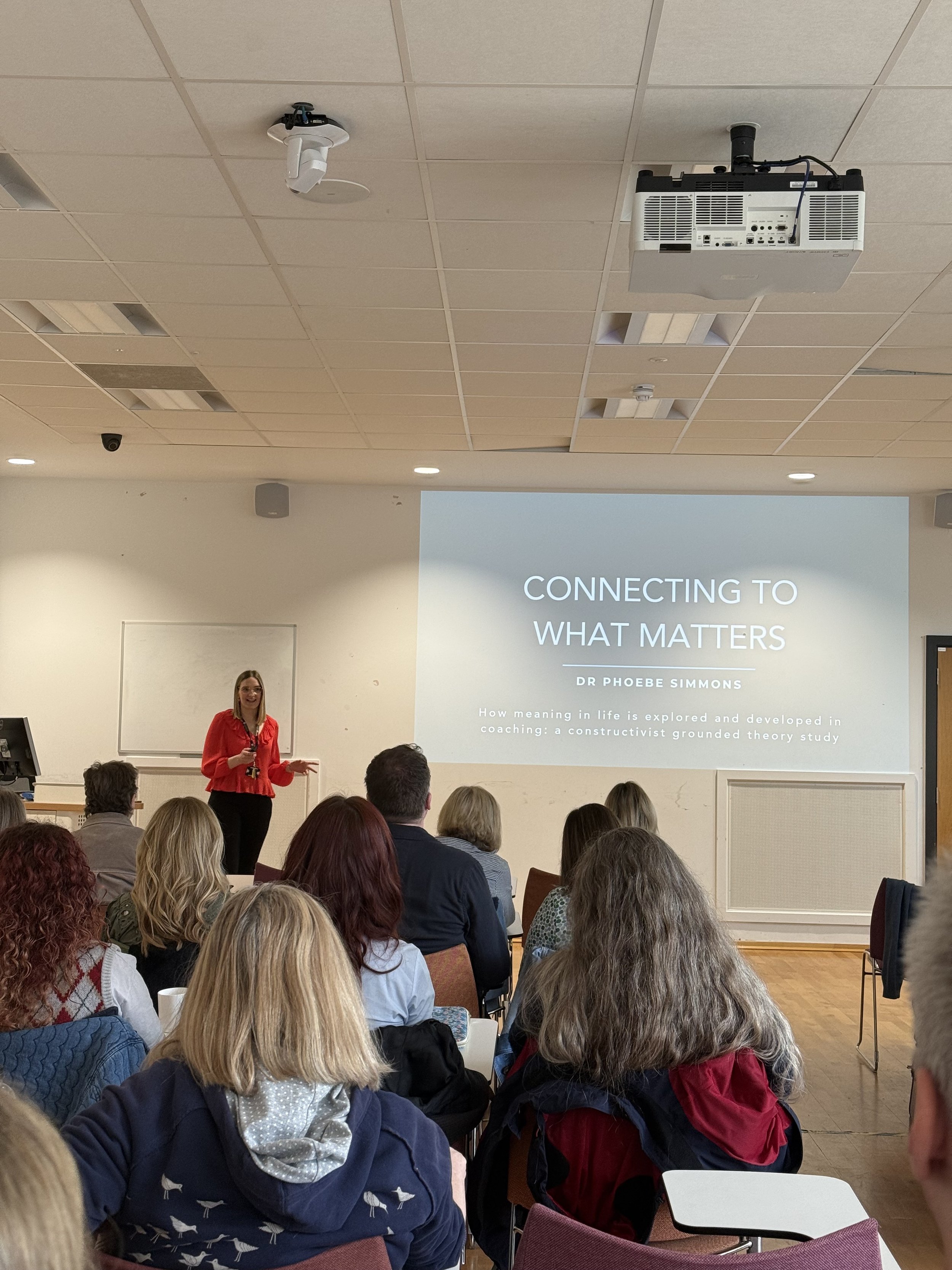 A woman in a red shirt giving a presentation in a conference room with a large screen displaying the title "Connecting to What Matters" and the speaker's name Dr. Phoebe Simmons, with an audience seated and listening.