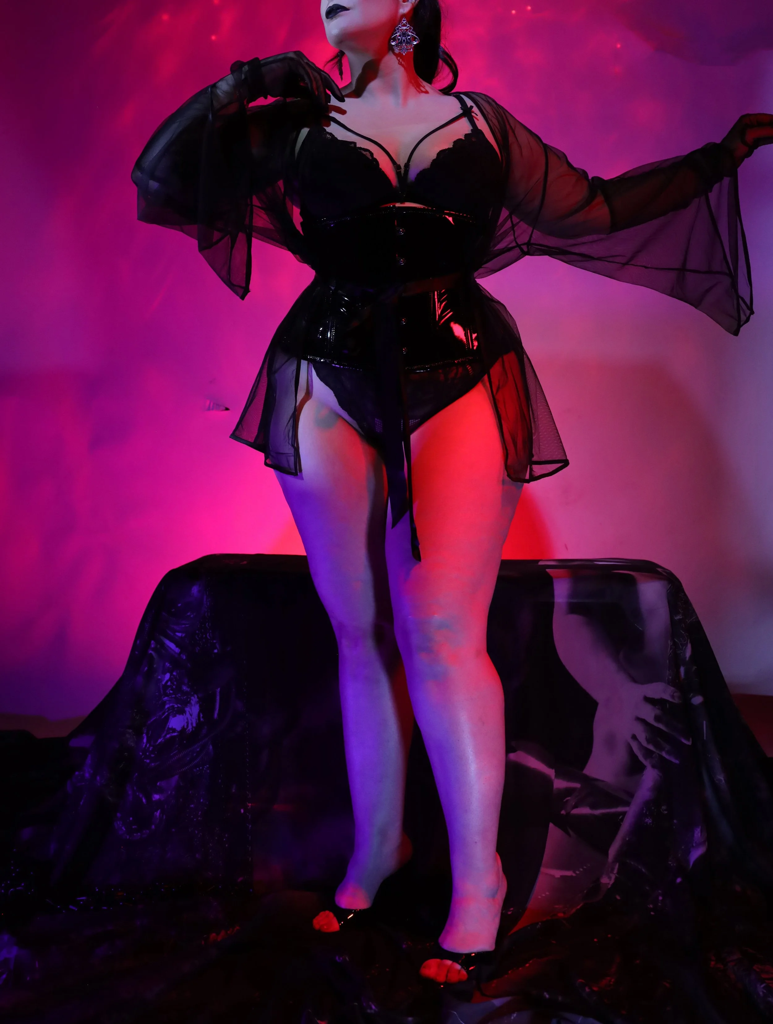 A woman dressed in black leather and sheer black fabric, posed with one arm elevated and the other resting on her shoulder, standing in front of a vibrant, neon pink and purple background.