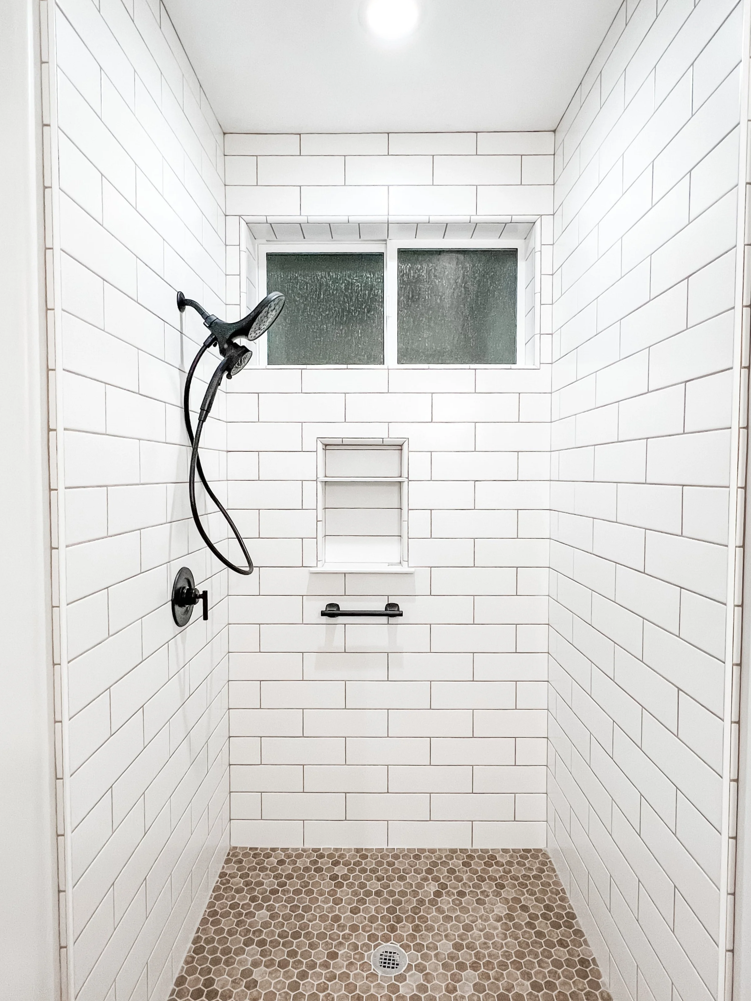 A walk-in shower with white subway tile walls, a small window, a black showerhead, and a brown hexagonal tile floor with a drain.