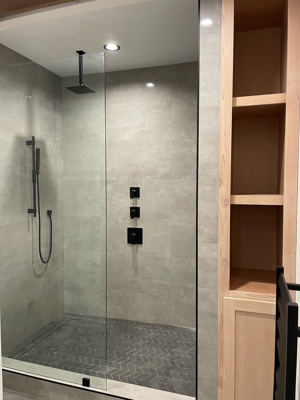 Modern shower with glass enclosure, rainfall showerhead, handheld shower, and black fixtures, adjacent to wooden shelving unit.