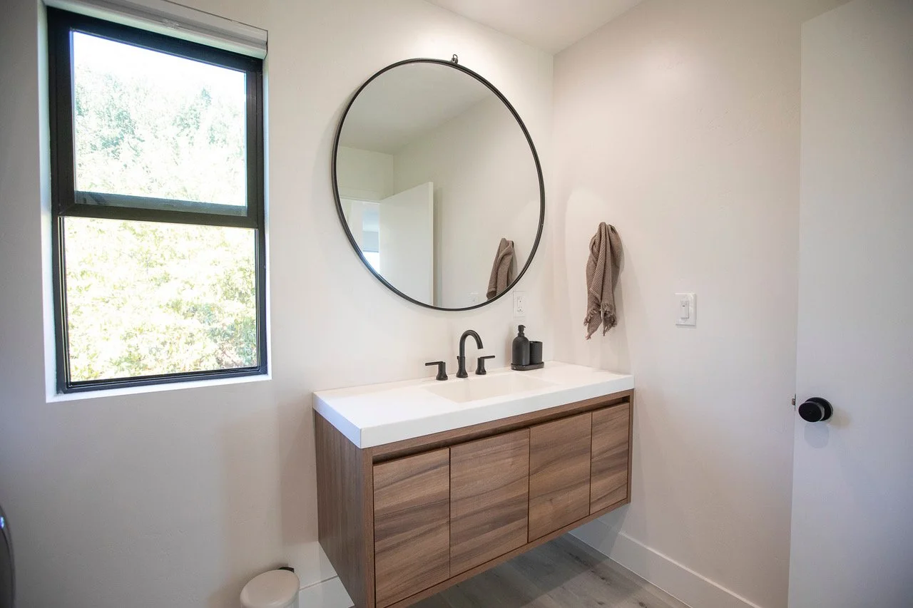 Modern bathroom with a wall-mounted wooden vanity, white countertop, black faucet, round mirror, window, and beige towels.