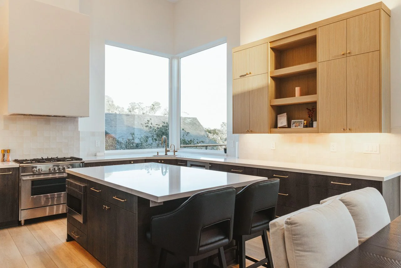 Modern kitchen with a large white island, black cabinets, and a stainless steel stove. Light wood upper cabinets and a window overlooking outdoor scenery. Dining chairs and part of a dining table are visible in the foreground.