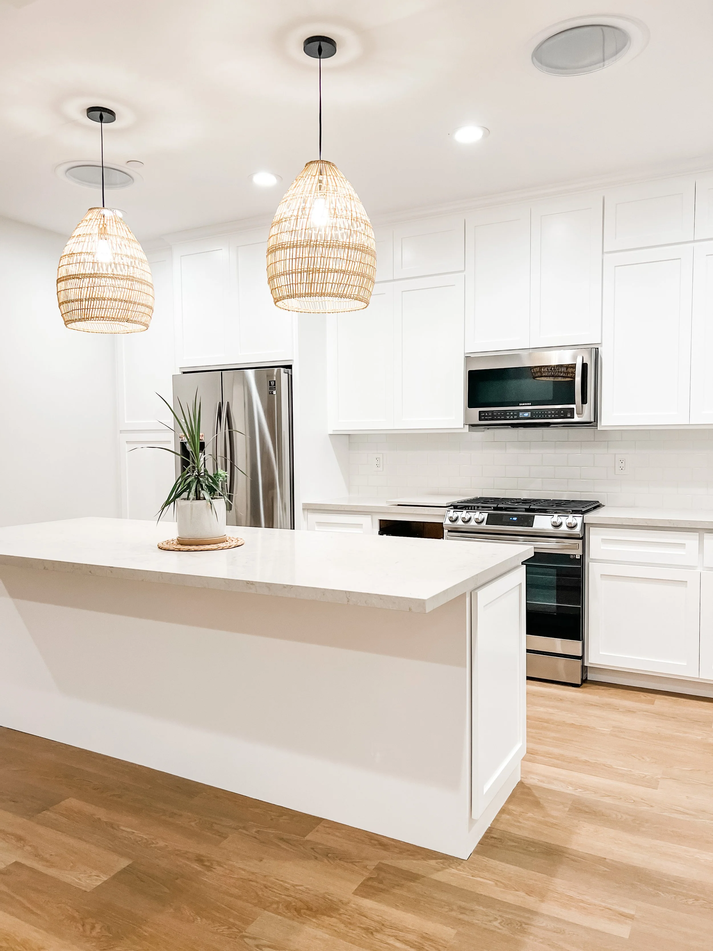 A modern kitchen with white cabinets, a white countertop island, stainless steel appliances, including a refrigerator, microwave, and stove, and wooden floors. Two wicker pendant lights hang above the island, and there is a potted plant on the island