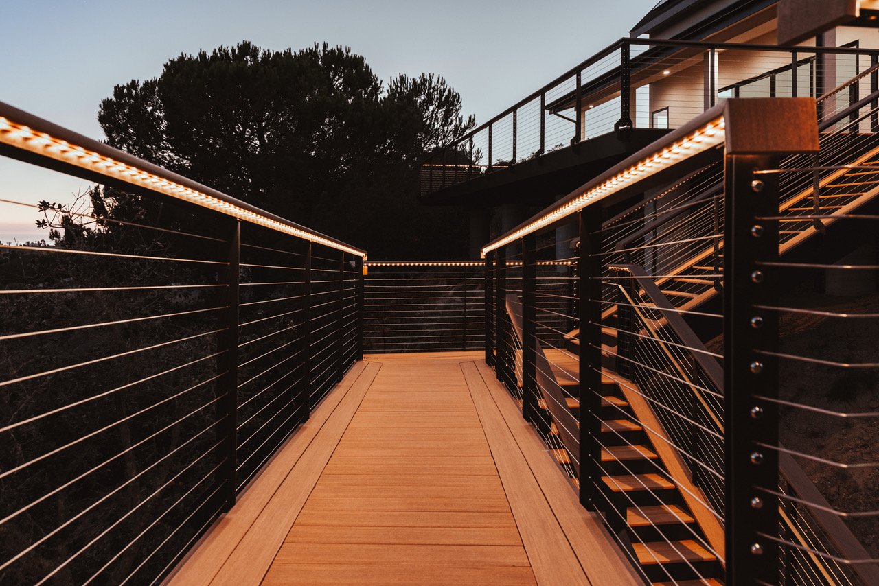 Modern outdoor balcony with wooden flooring, metal railing with horizontal wires, and attached staircase, illuminated by LED strip lighting, overlooking trees at dusk.