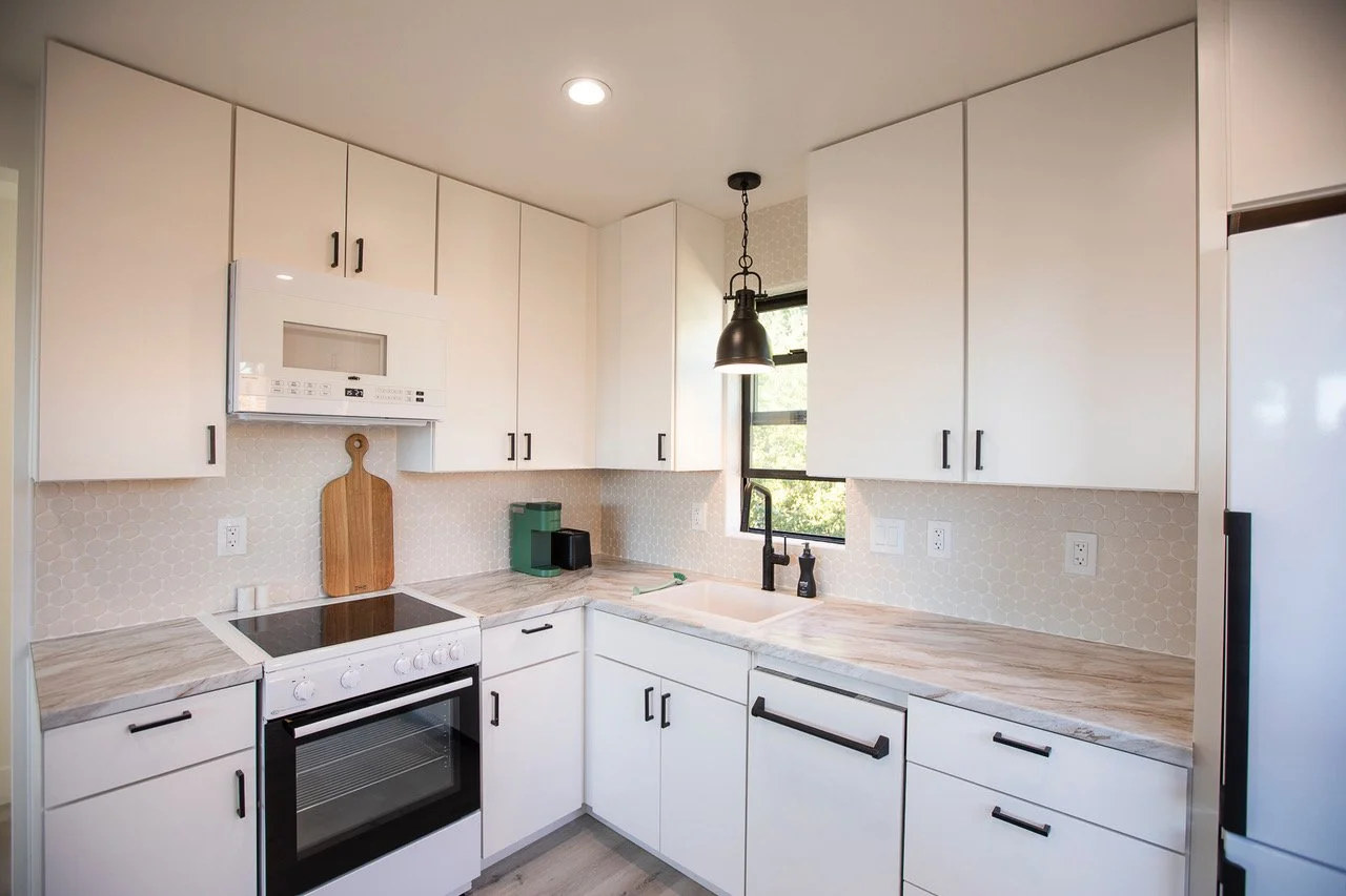 Modern white kitchen with black handles, marble countertops, electric stove, microwave, black faucet, and window with greenery outside
